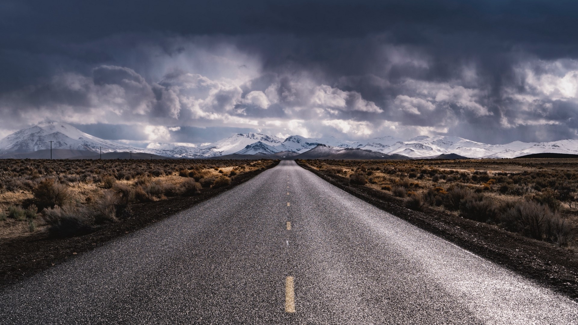 Empty road leading to snow-capped mountains under stormy clouds HD Wallpaper