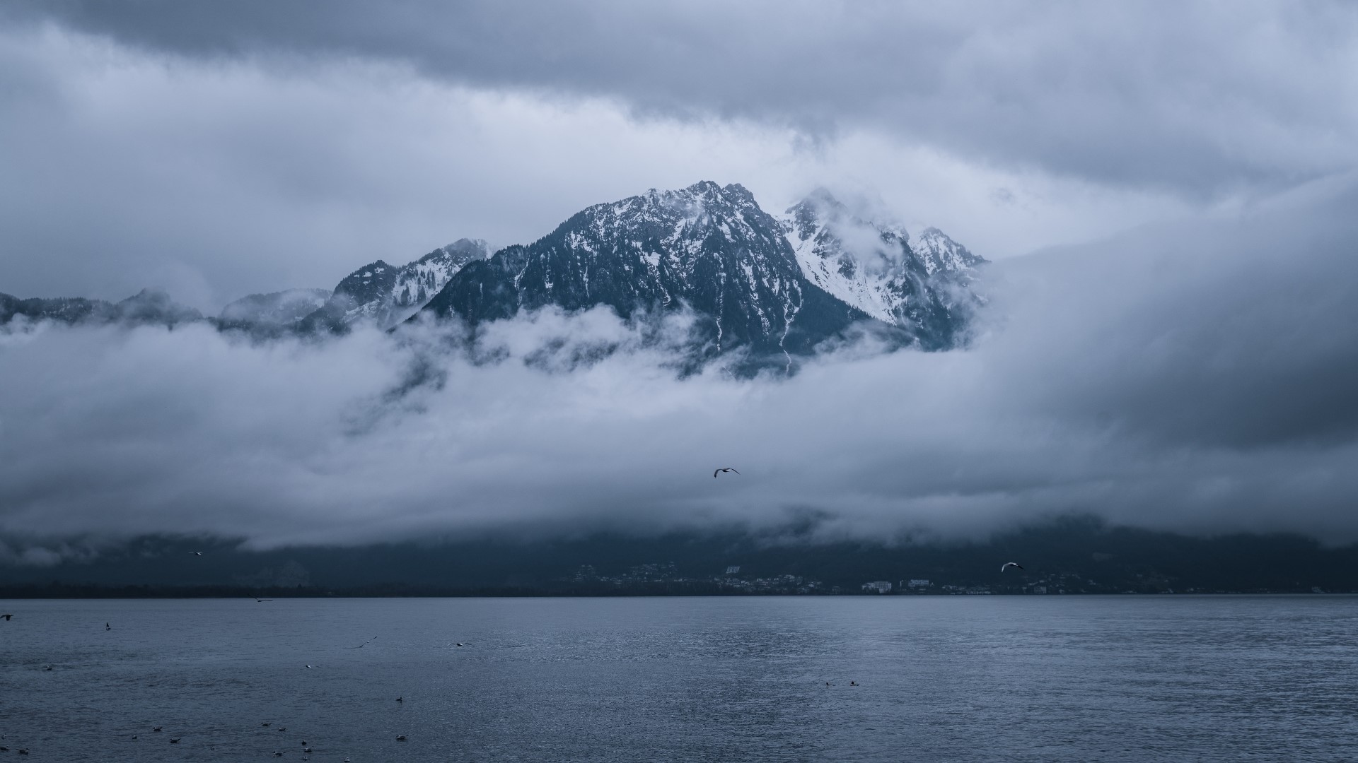 Snow-capped mountains shrouded in clouds over a lake HD Wallpaper