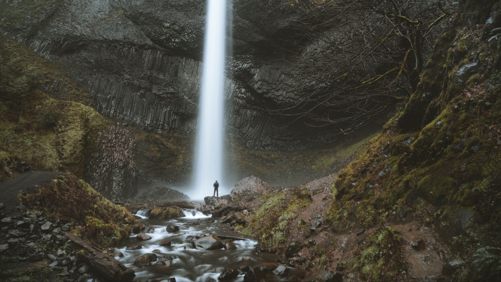Person standing at the base of a tall waterfall HD Wallpaper