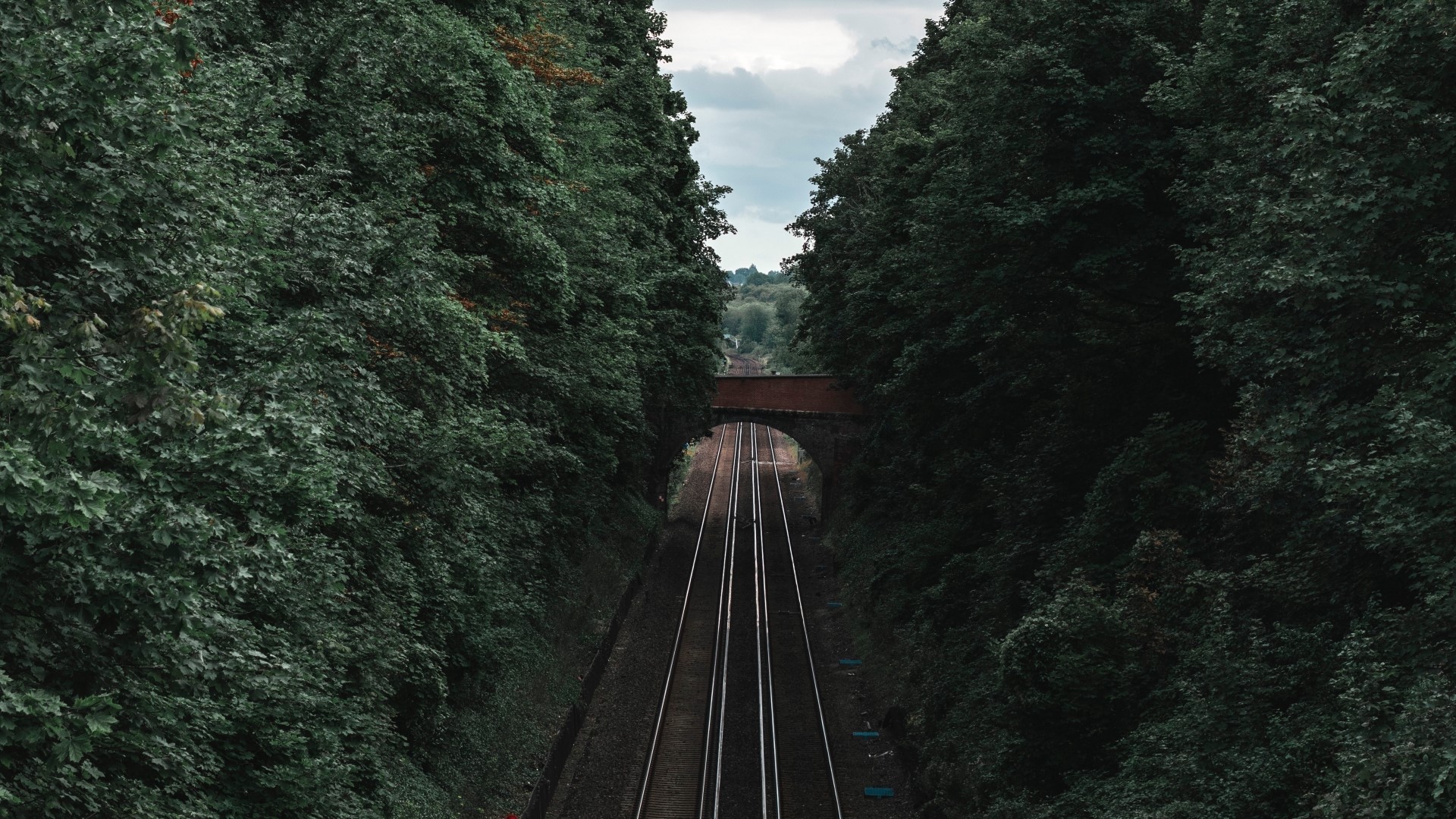 Railway tracks passing under a brick bridge surrounded by trees HD Wallpaper