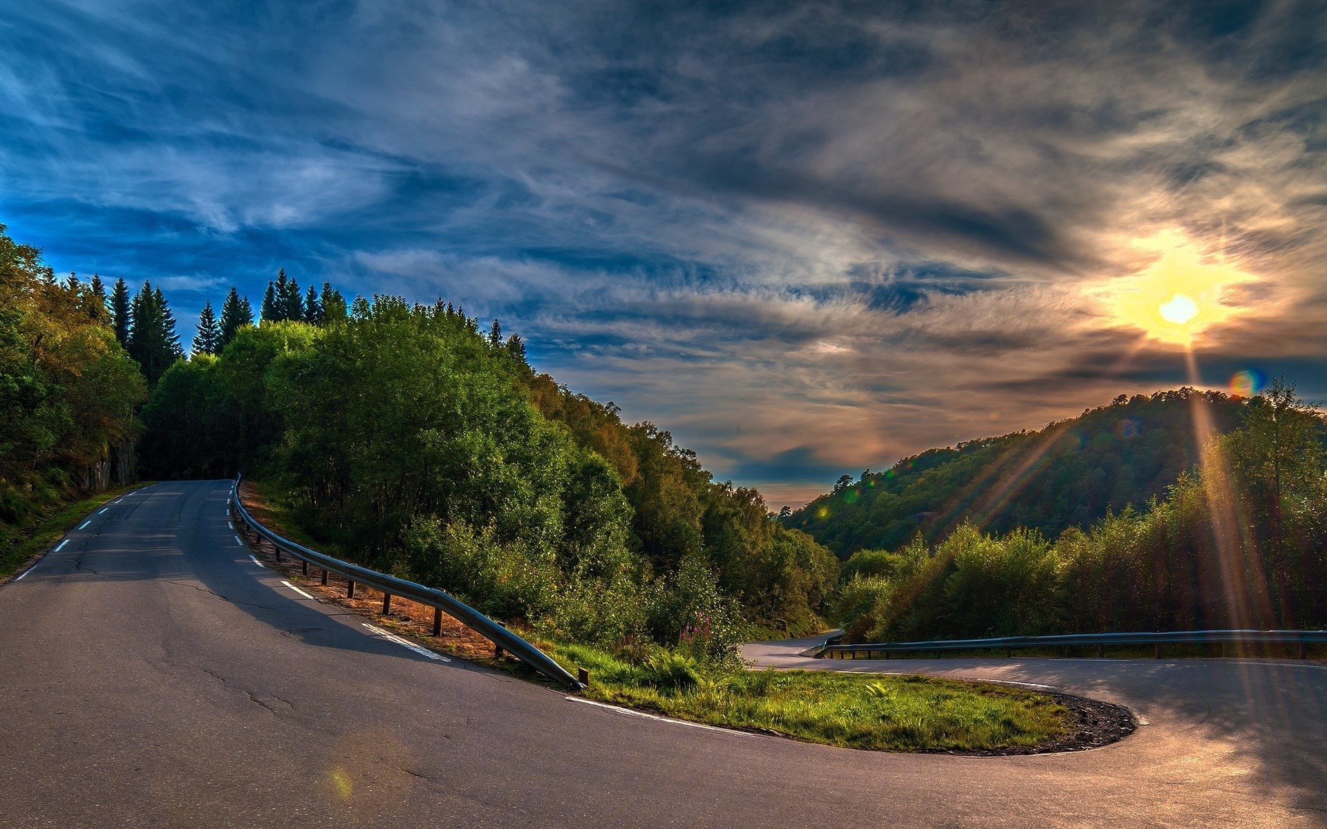 Winding mountain road with sunlit sky and trees HD Wallpaper