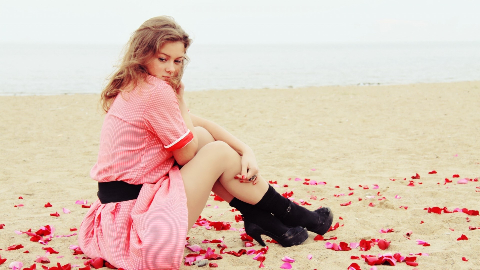 Woman in pink dress sitting on beach with rose petals HD Wallpaper