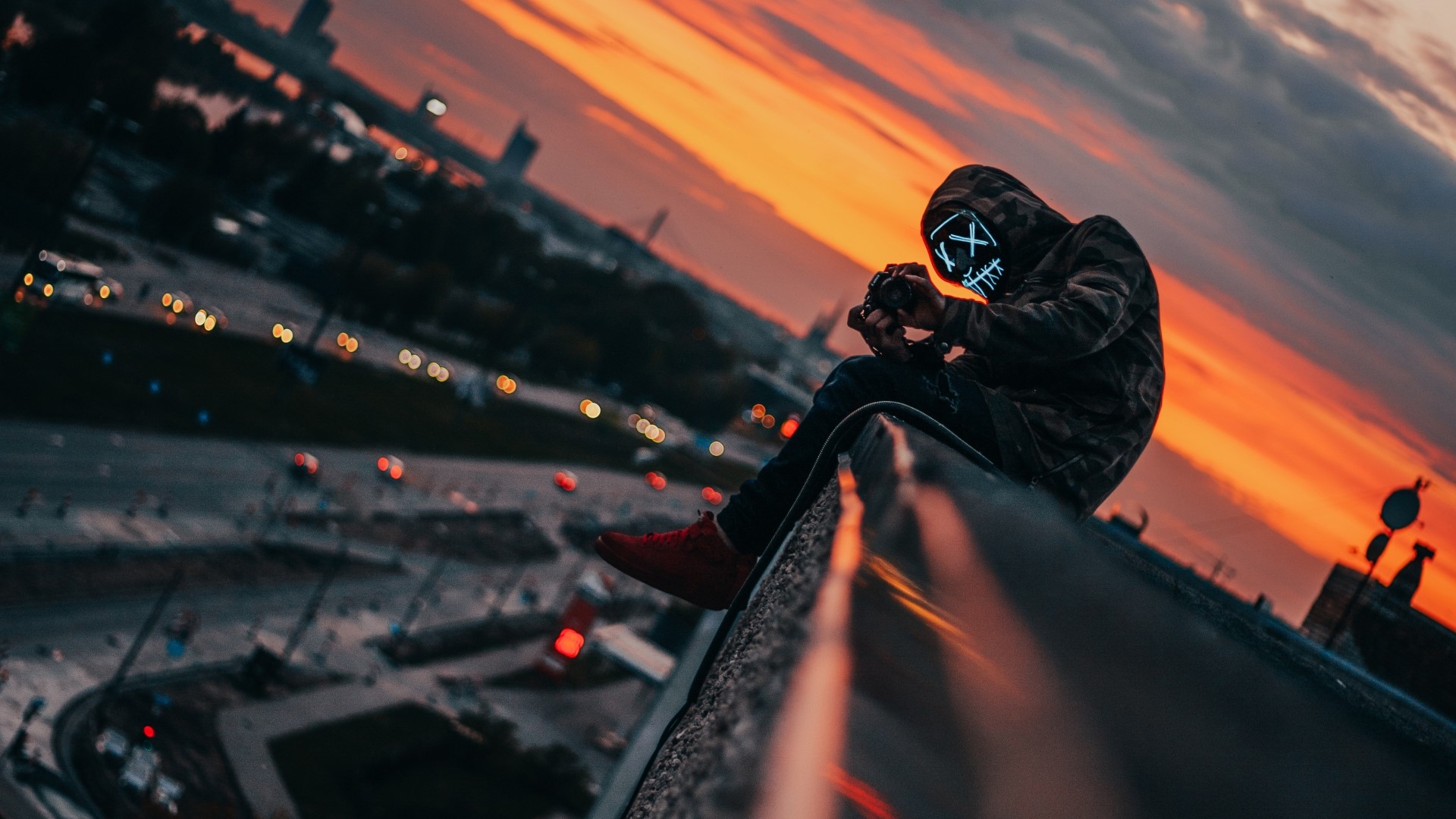 Person in mask sitting on edge of building at sunset HD Wallpaper