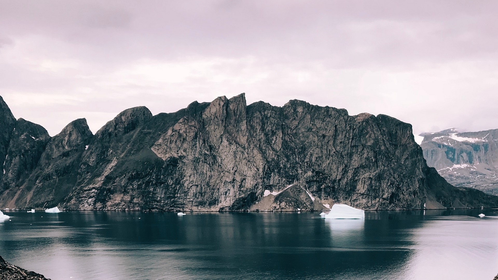 Jagged mountains rise from a dark, reflective body of water with icebergs. HD Wallpaper