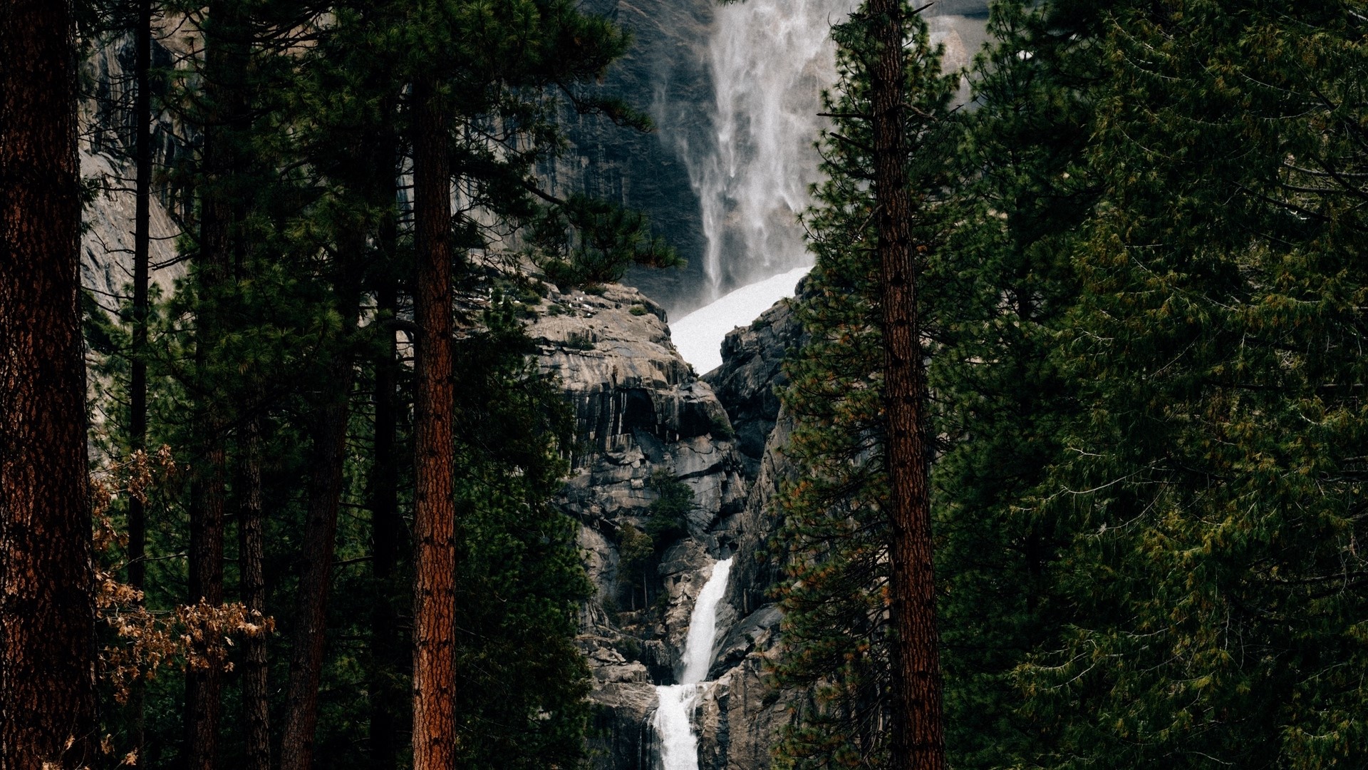 Waterfall cascading down rocky cliffs surrounded by pine trees HD Wallpaper