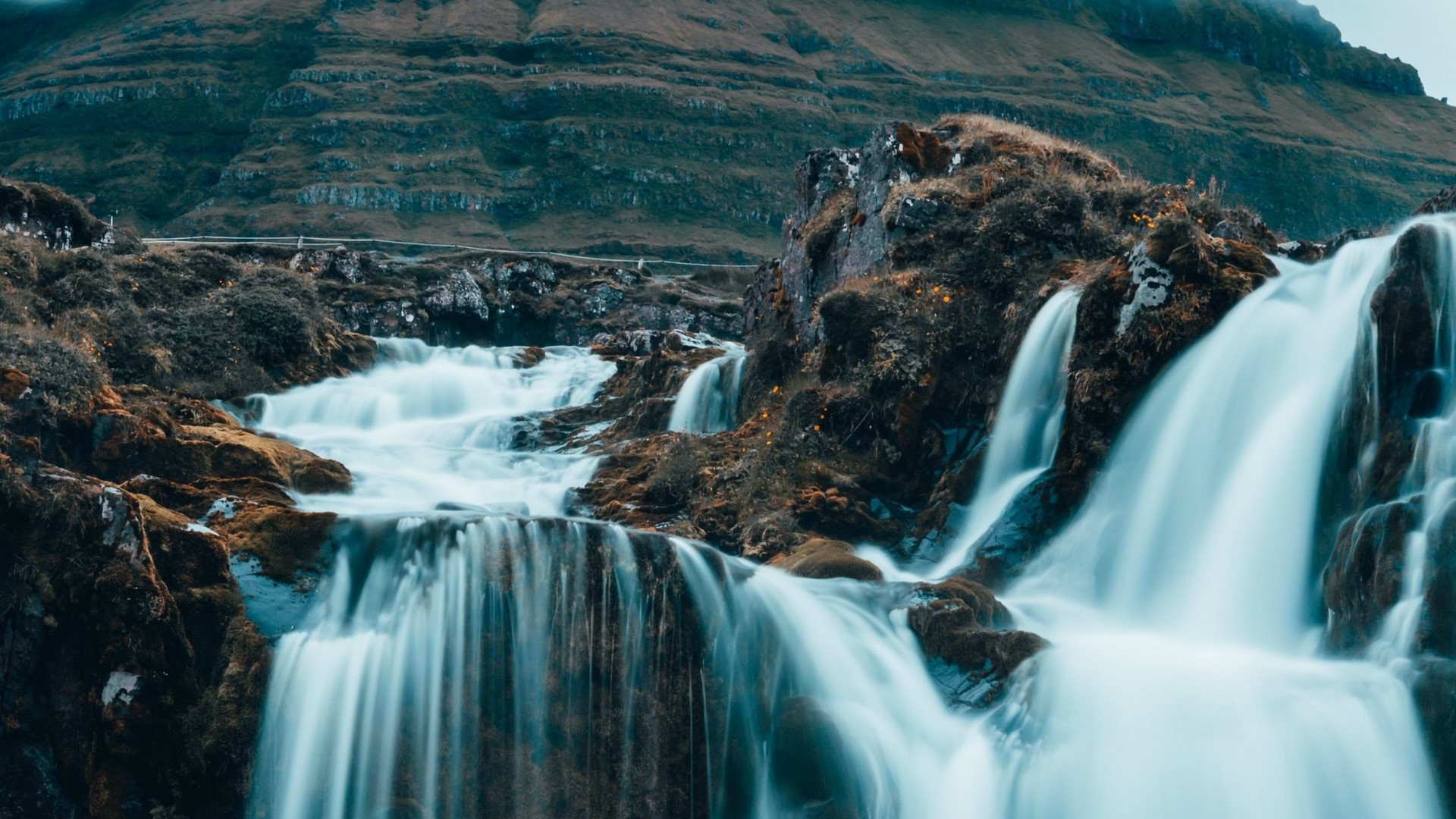 Cascading waterfalls over mossy rocks with a mountain backdrop HD Wallpaper