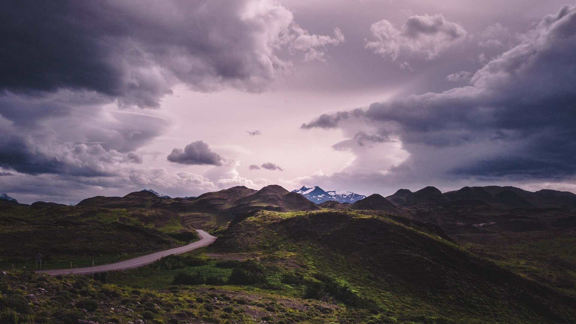 Road through rolling hills towards snow-capped mountains under stormy clouds HD Wallpaper