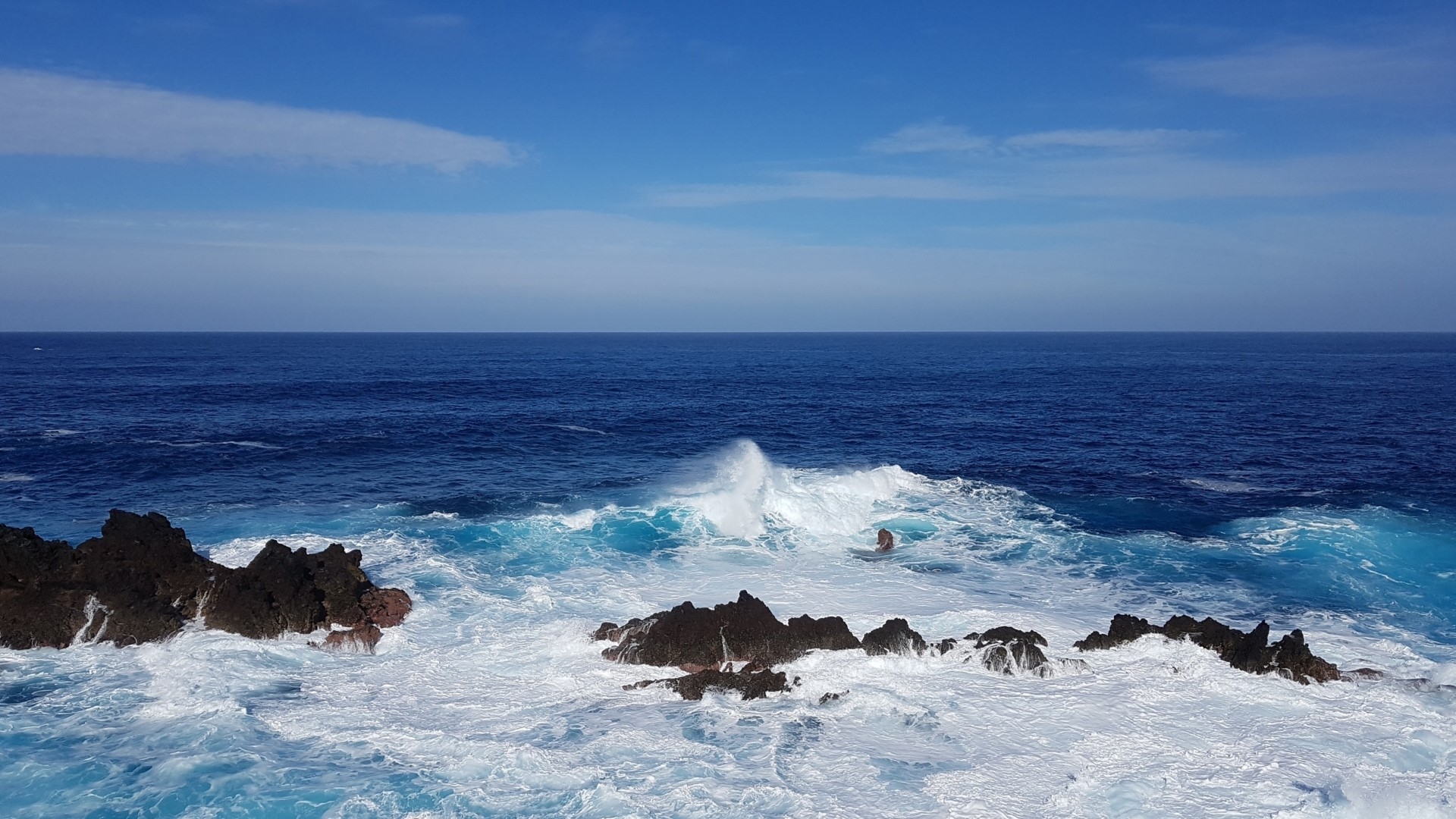 Ocean waves crashing on rocks under a blue sky HD Wallpaper