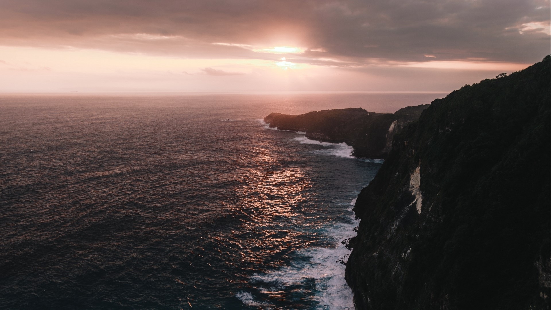 Ocean waves crash against a cliffside at sunset HD Wallpaper