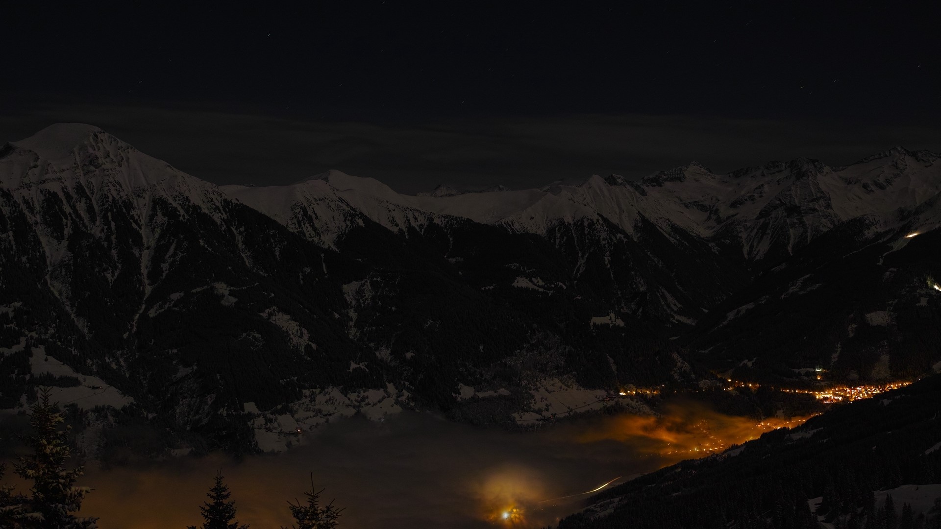 Snowy mountains at night with a village below HD Wallpaper