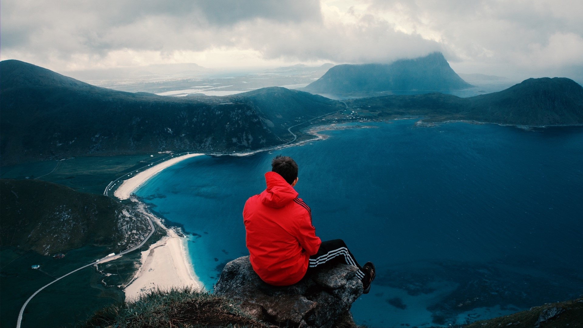 Man in red jacket sitting on cliff overlooking ocean and mountains HD Wallpaper