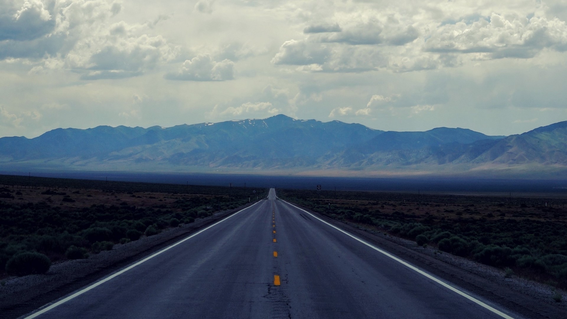 Long empty road leading to mountains under cloudy sky HD Wallpaper
