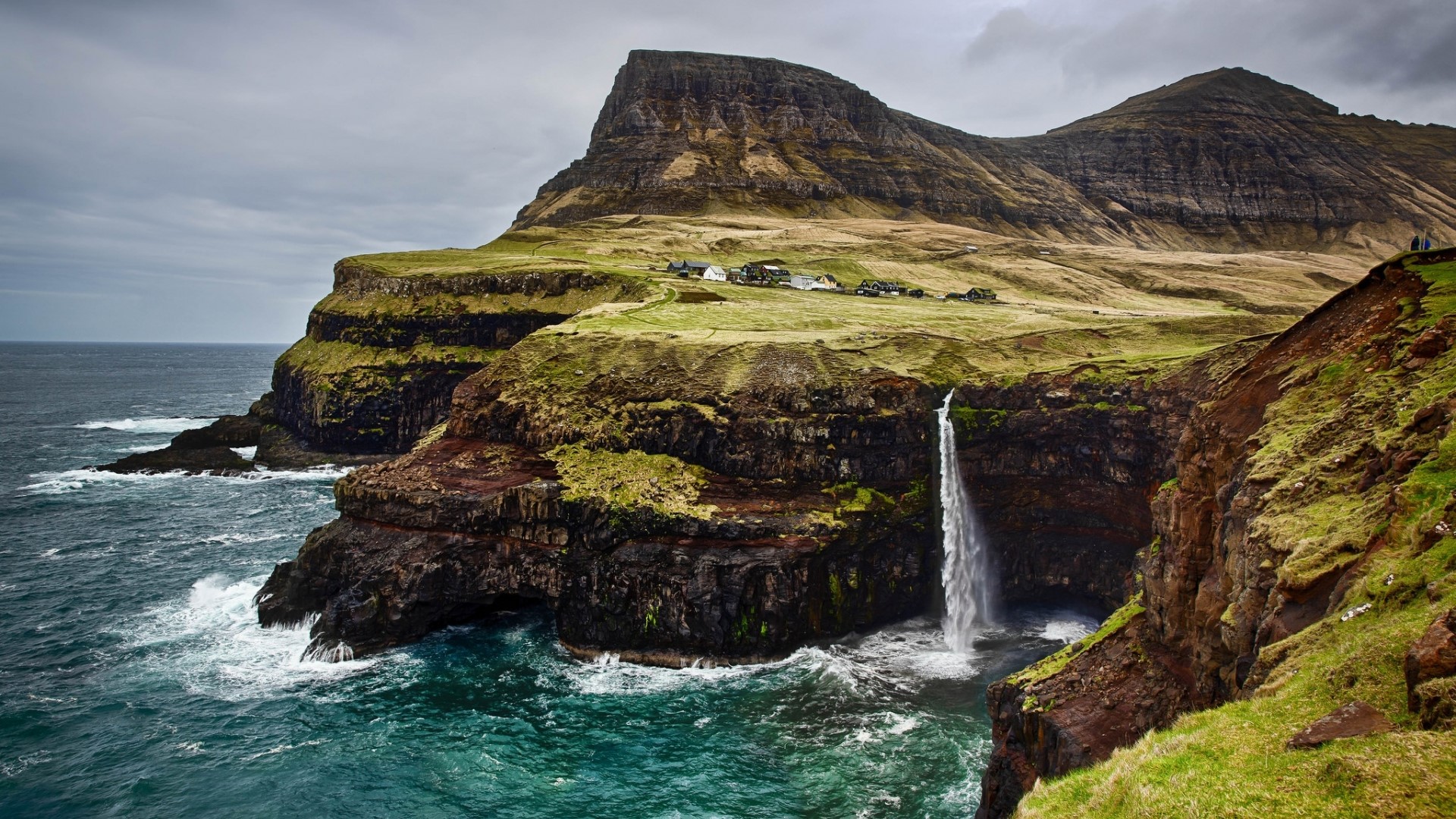 Waterfall cascading into the ocean near a village in the Faroe Islands HD Wallpaper