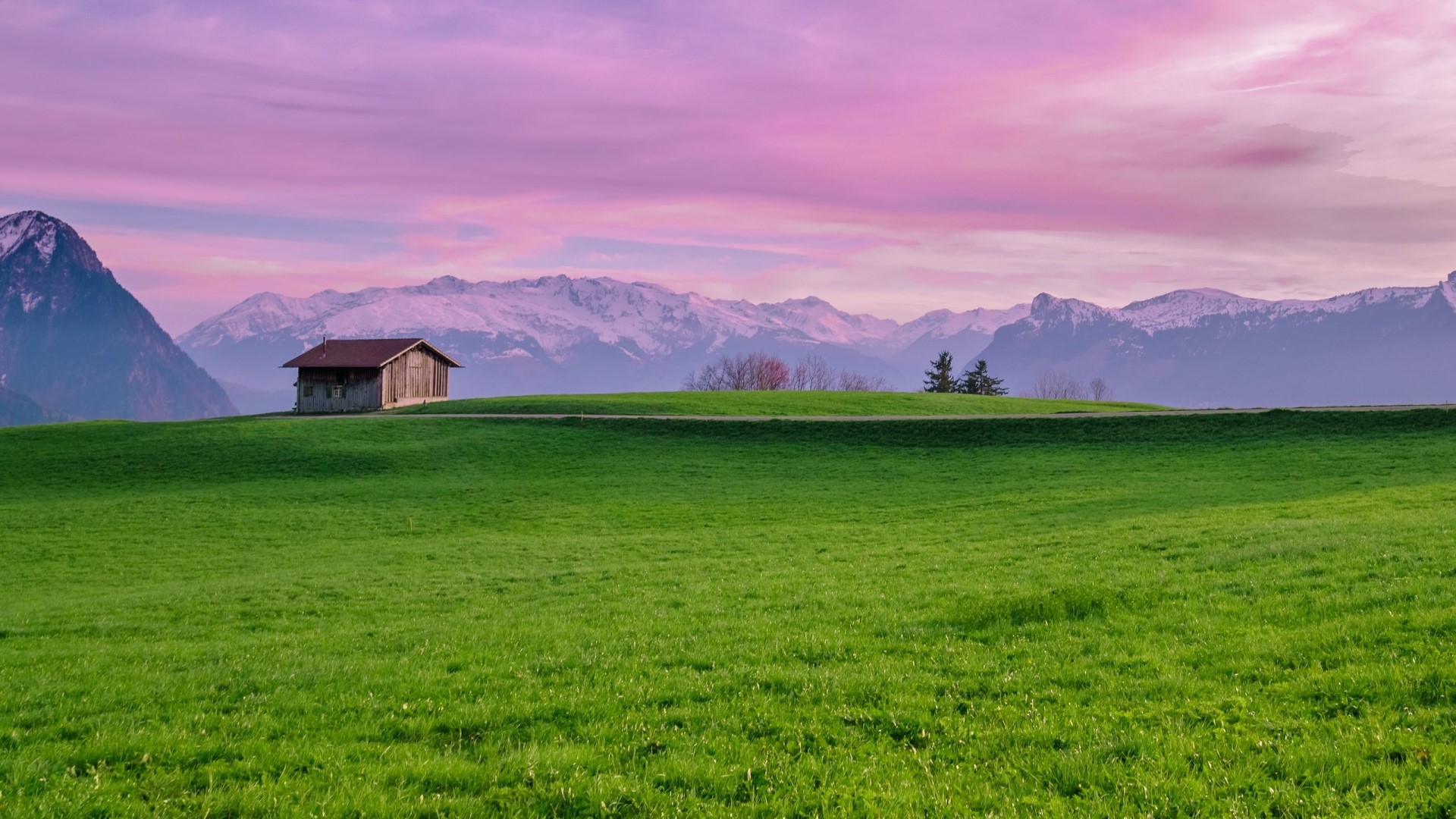 Wooden cabin on a grassy hill with snow-capped mountains and pink sky HD Wallpaper