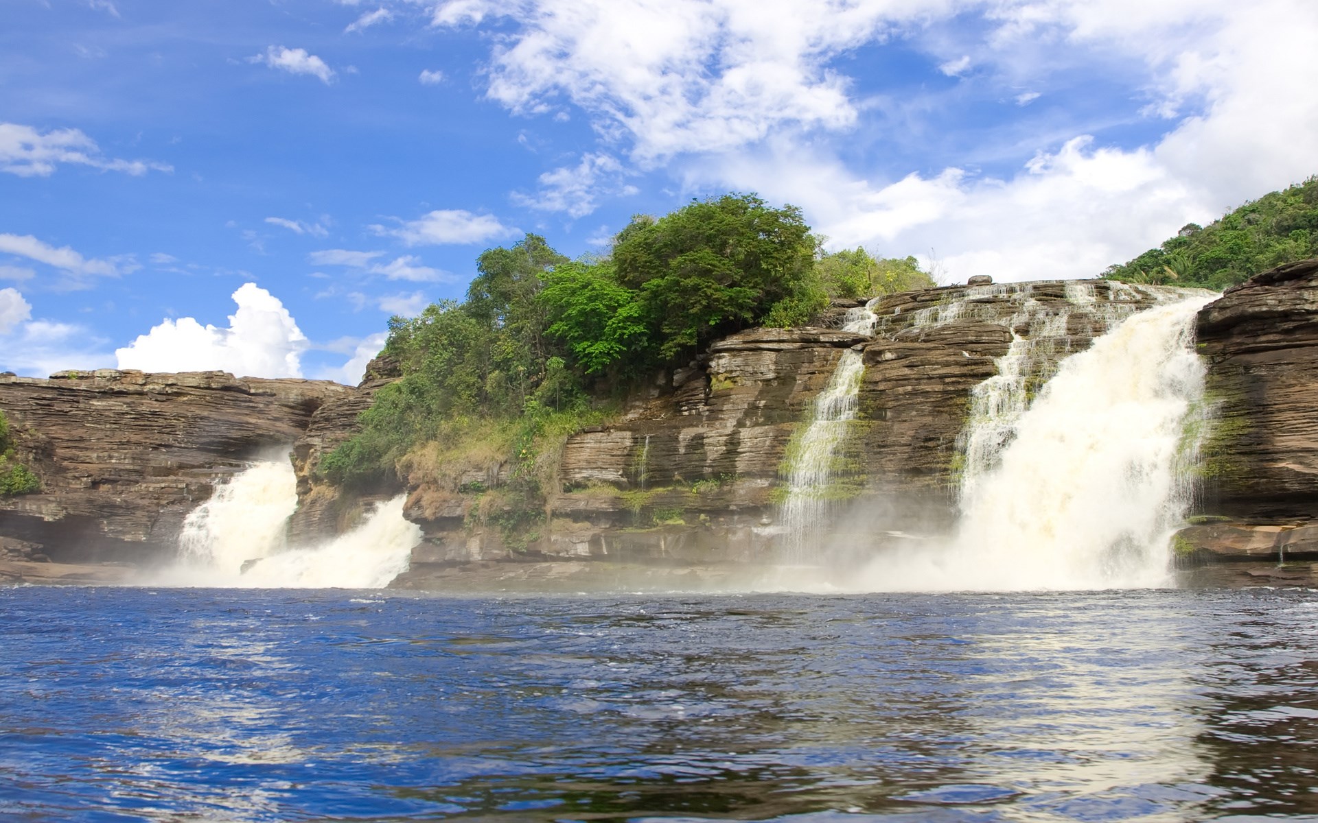 Waterfalls cascading over rocky cliffs into a blue river HD Wallpaper