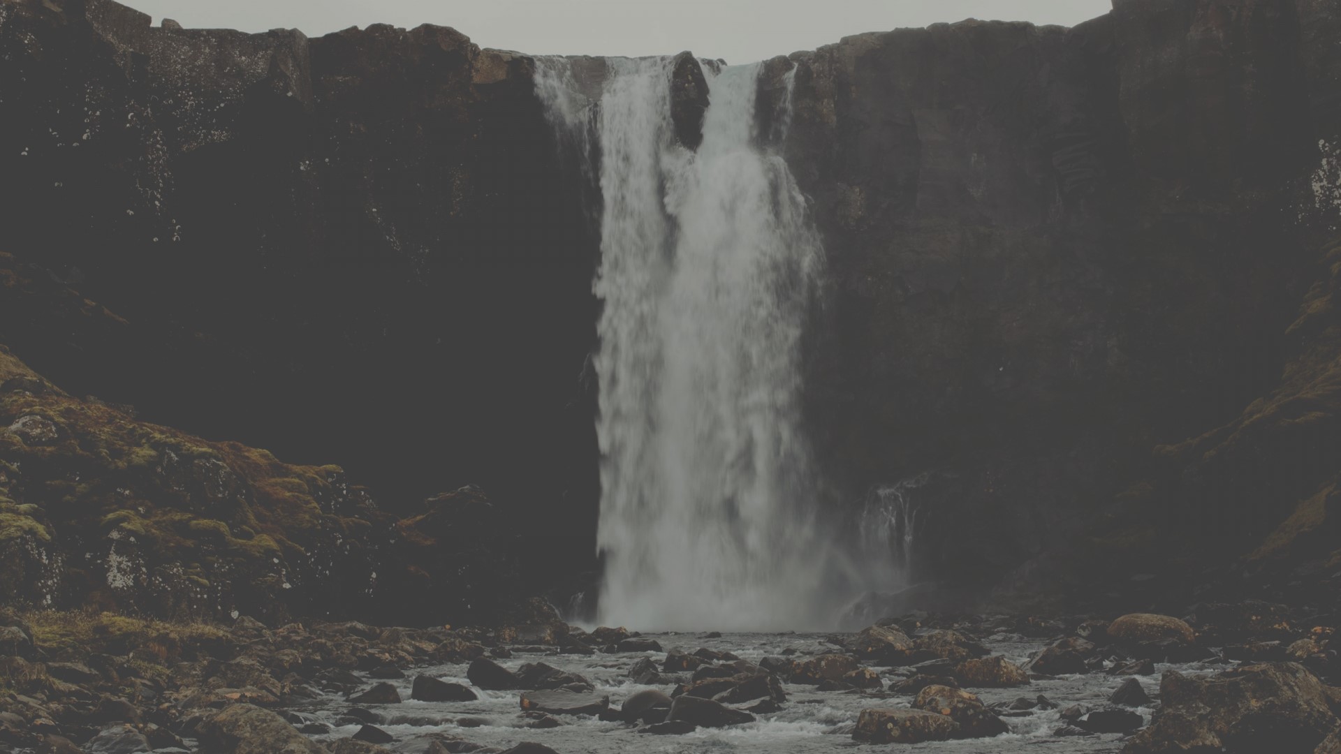Waterfall cascading over dark cliffs into a rocky riverbed HD Wallpaper
