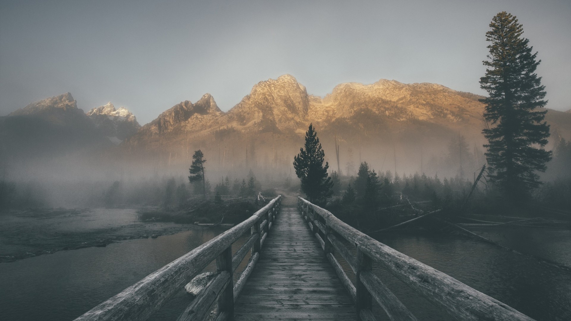 Wooden bridge over a misty river with mountains HD Wallpaper
