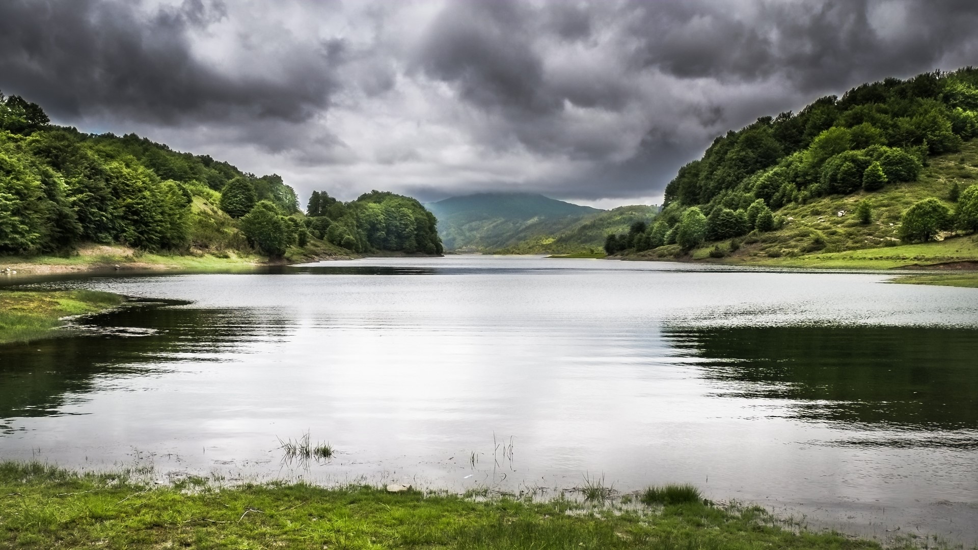 Lake surrounded by green hills under cloudy sky HD Wallpaper