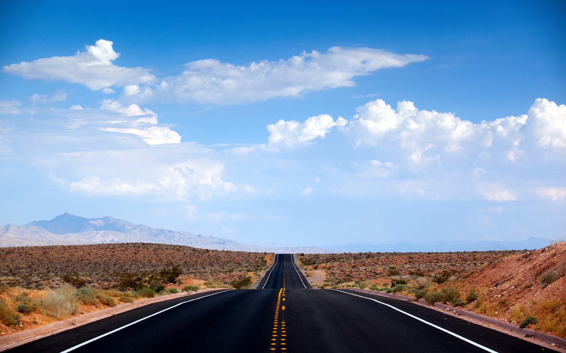 Desert highway stretching towards mountains under a cloudy sky HD Wallpaper