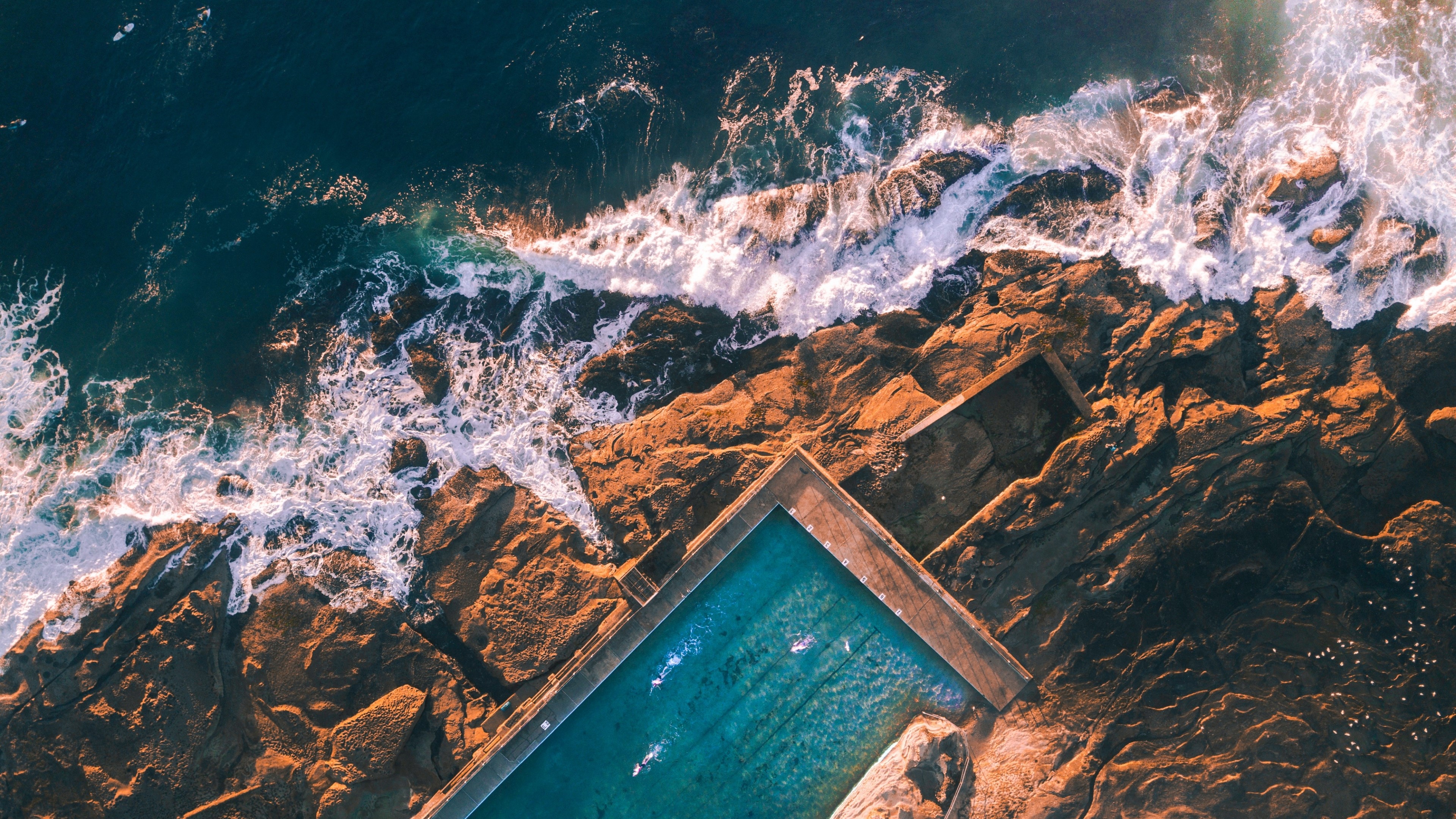 Aerial view of ocean waves crashing on rocks near a swimming pool HD Wallpaper