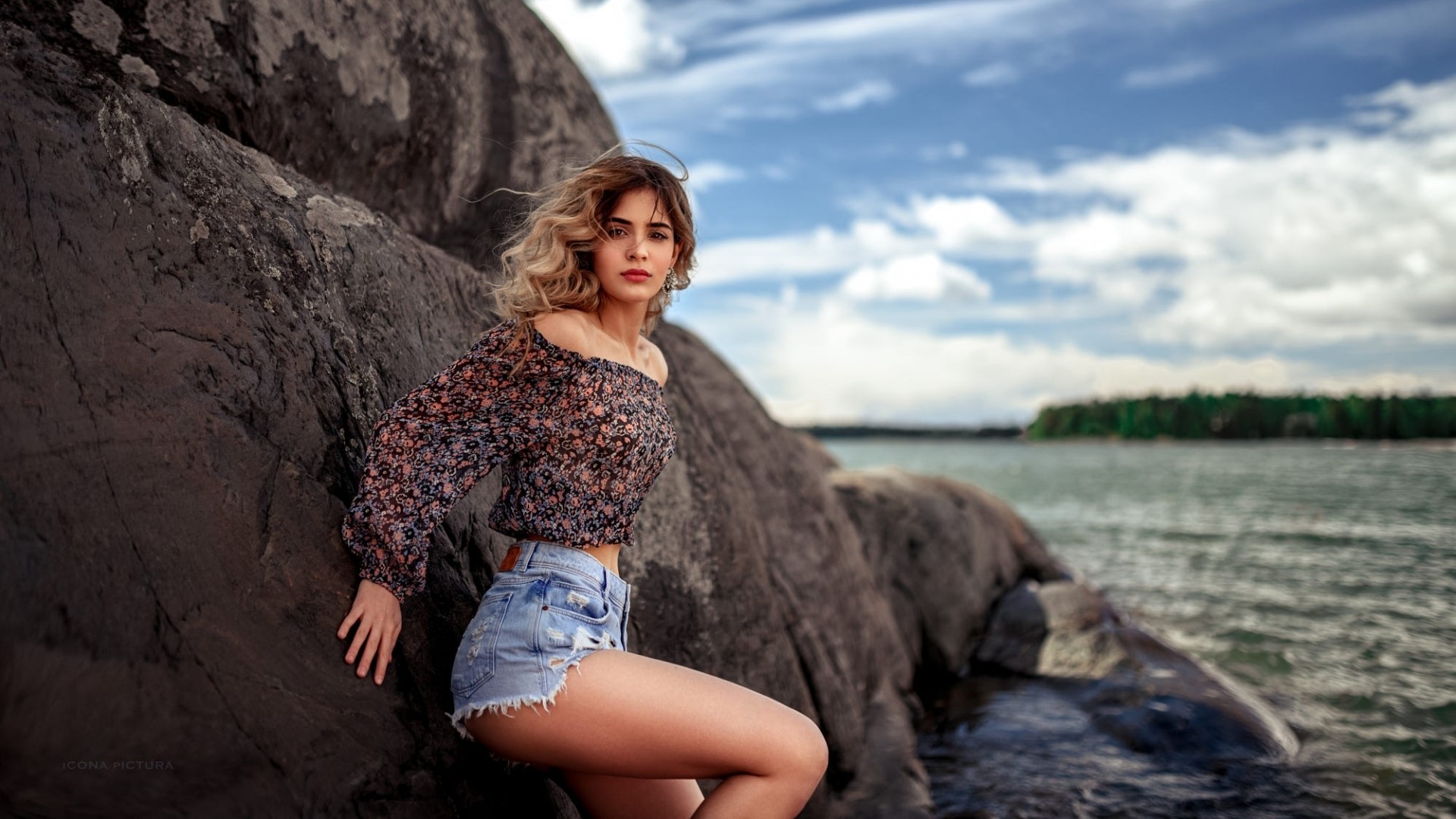 Woman in shorts and off-the-shoulder top on rocks by the water HD Wallpaper