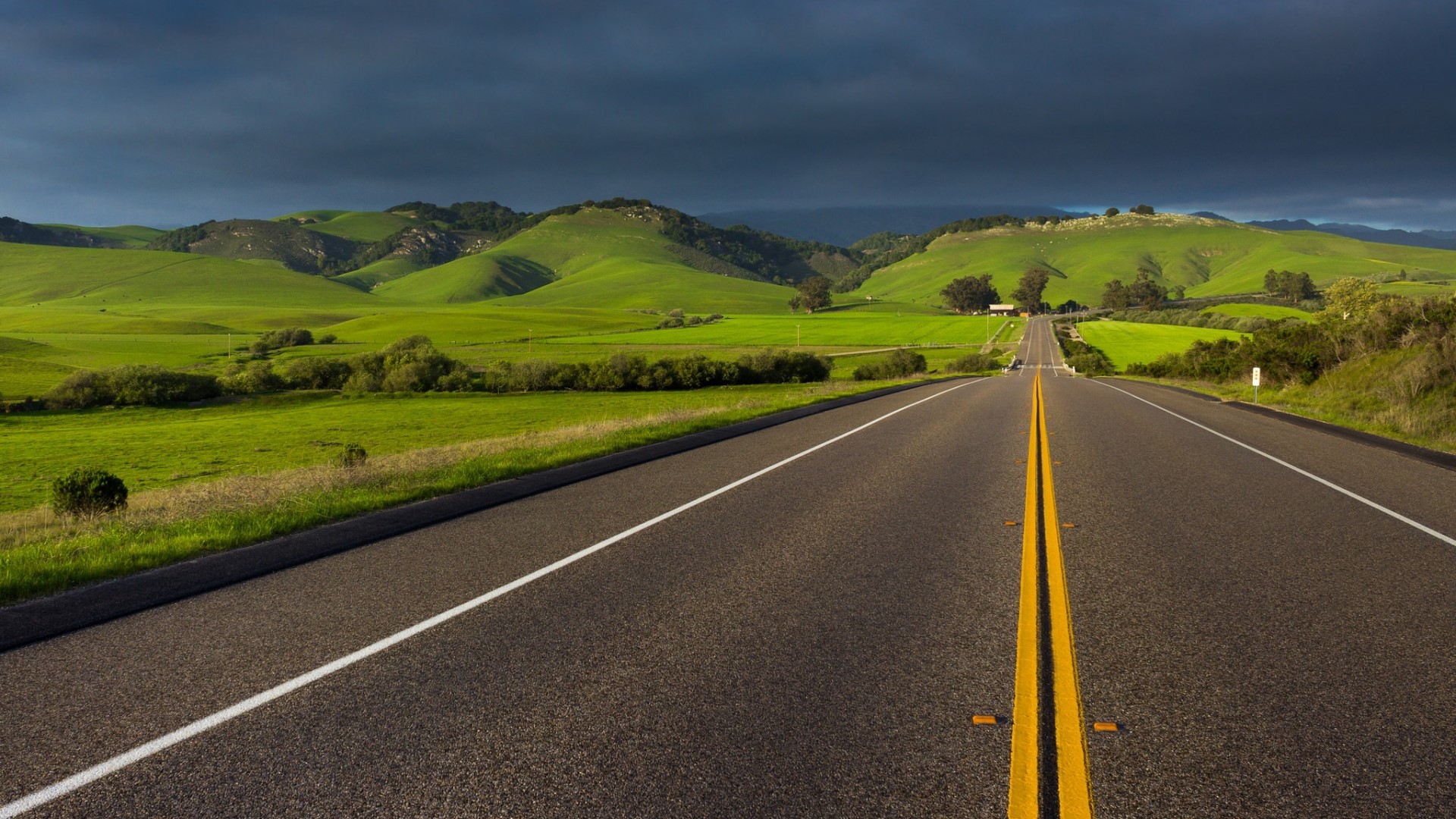 Road through green rolling hills under dark clouds HD Wallpaper