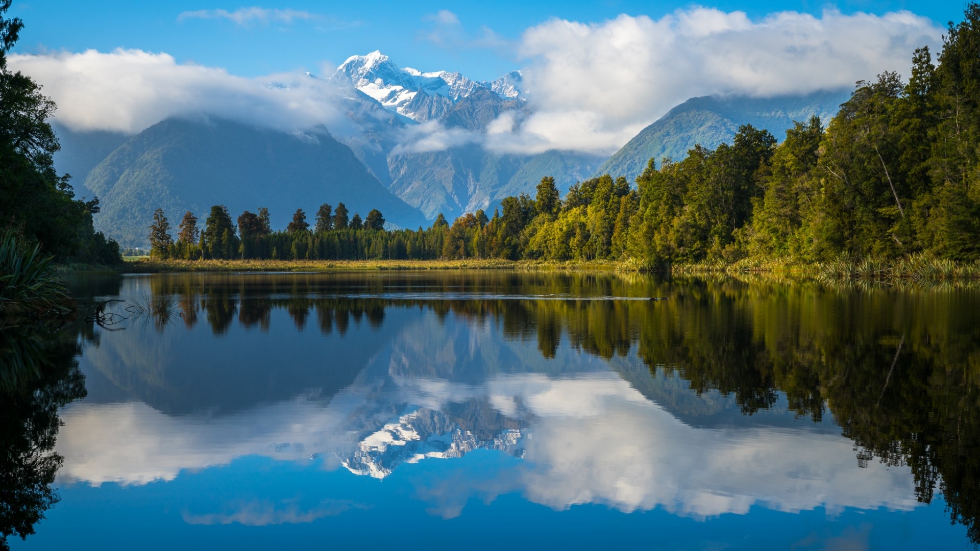 Snow-capped mountains reflected in a calm lake with trees HD Wallpaper