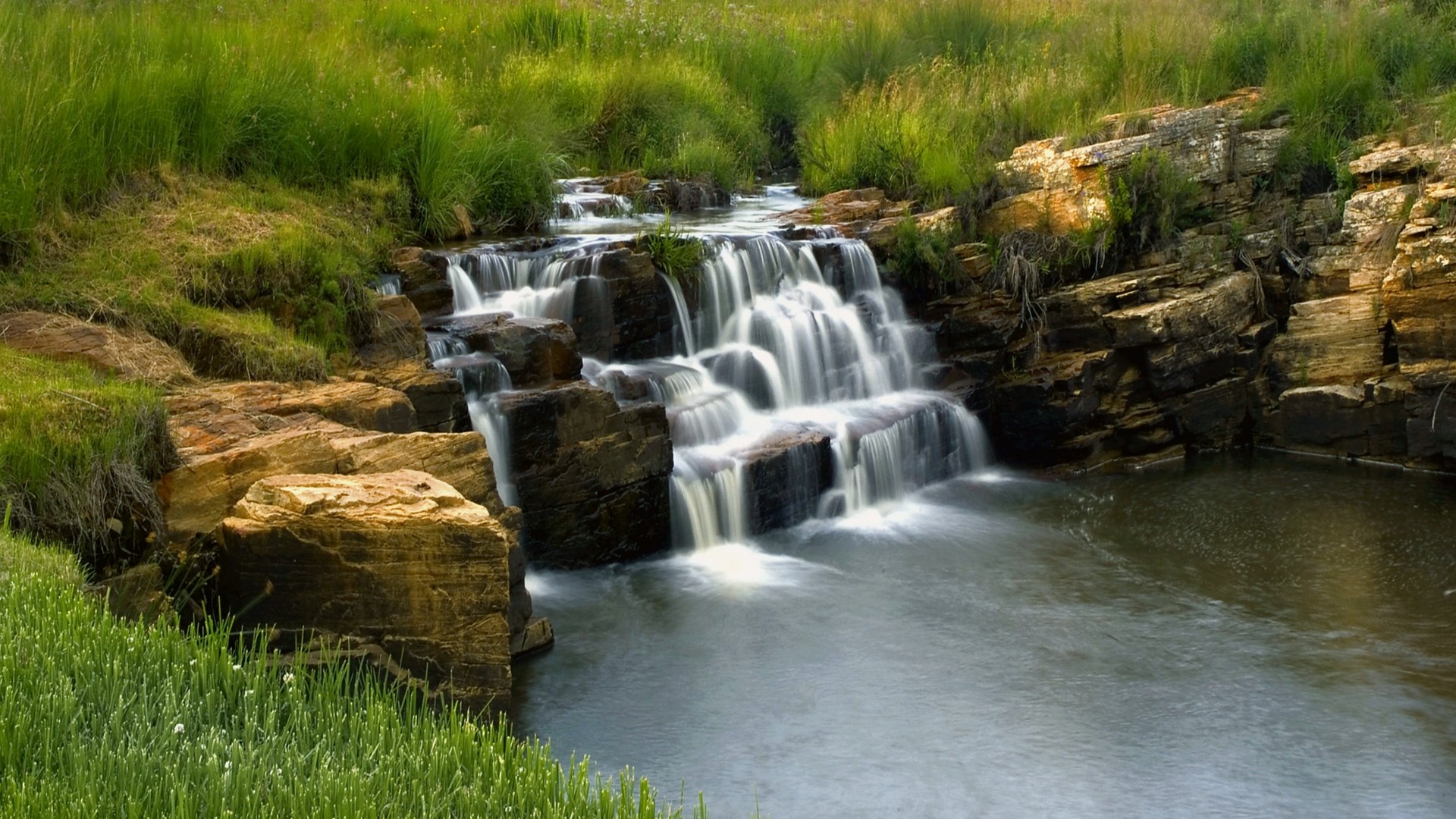 Waterfall cascading over rocks in a grassy landscape HD Wallpaper