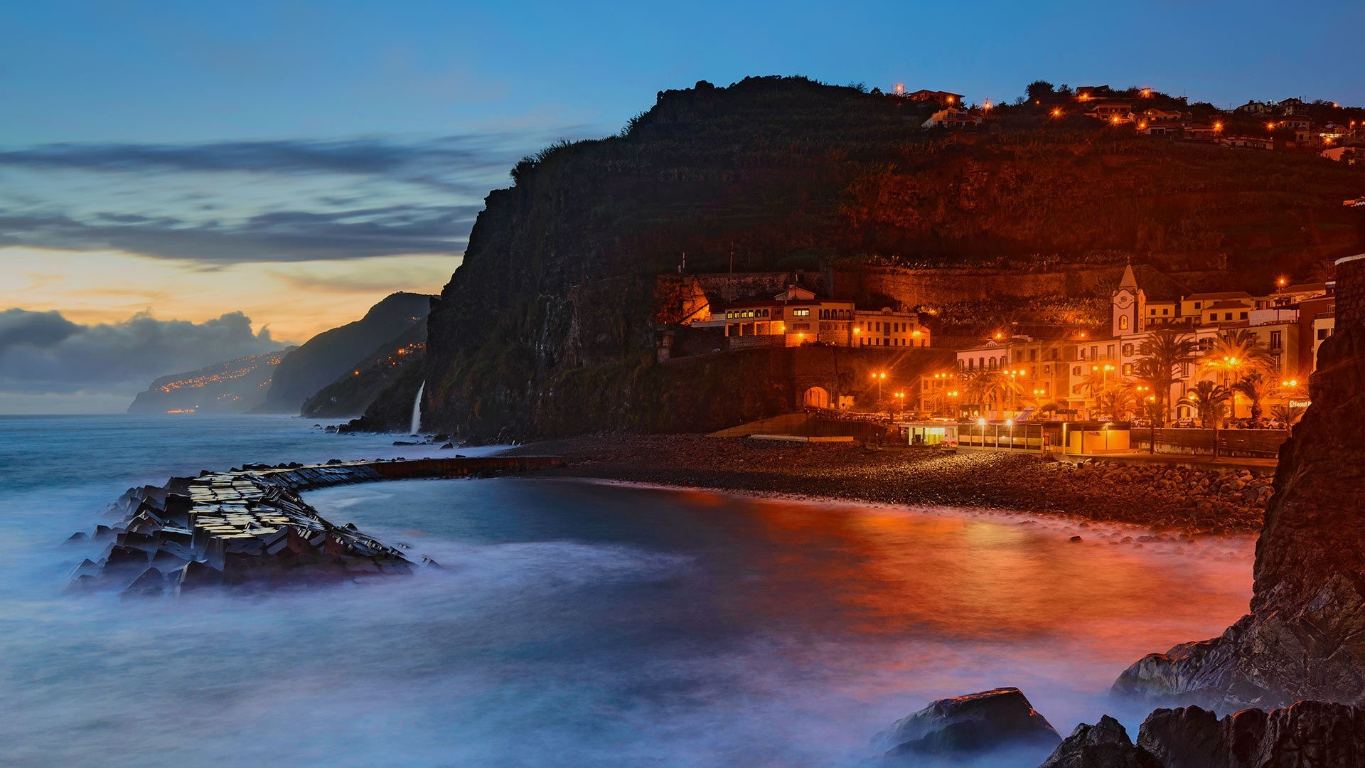 Madeira coastal town at dusk with illuminated buildings and ocean waves HD Wallpaper
