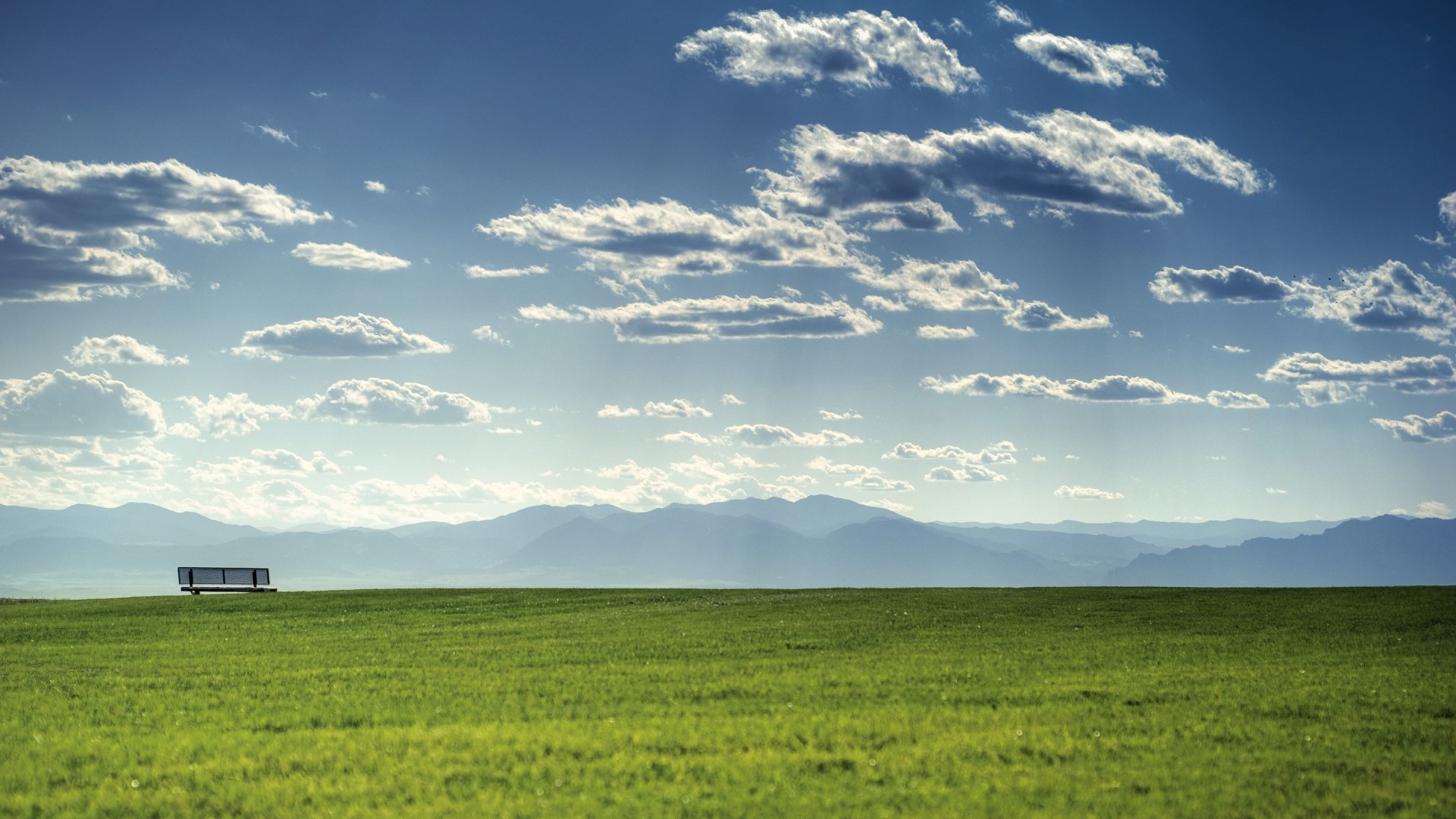 Empty bench on a grassy field with mountains and clouds HD Wallpaper