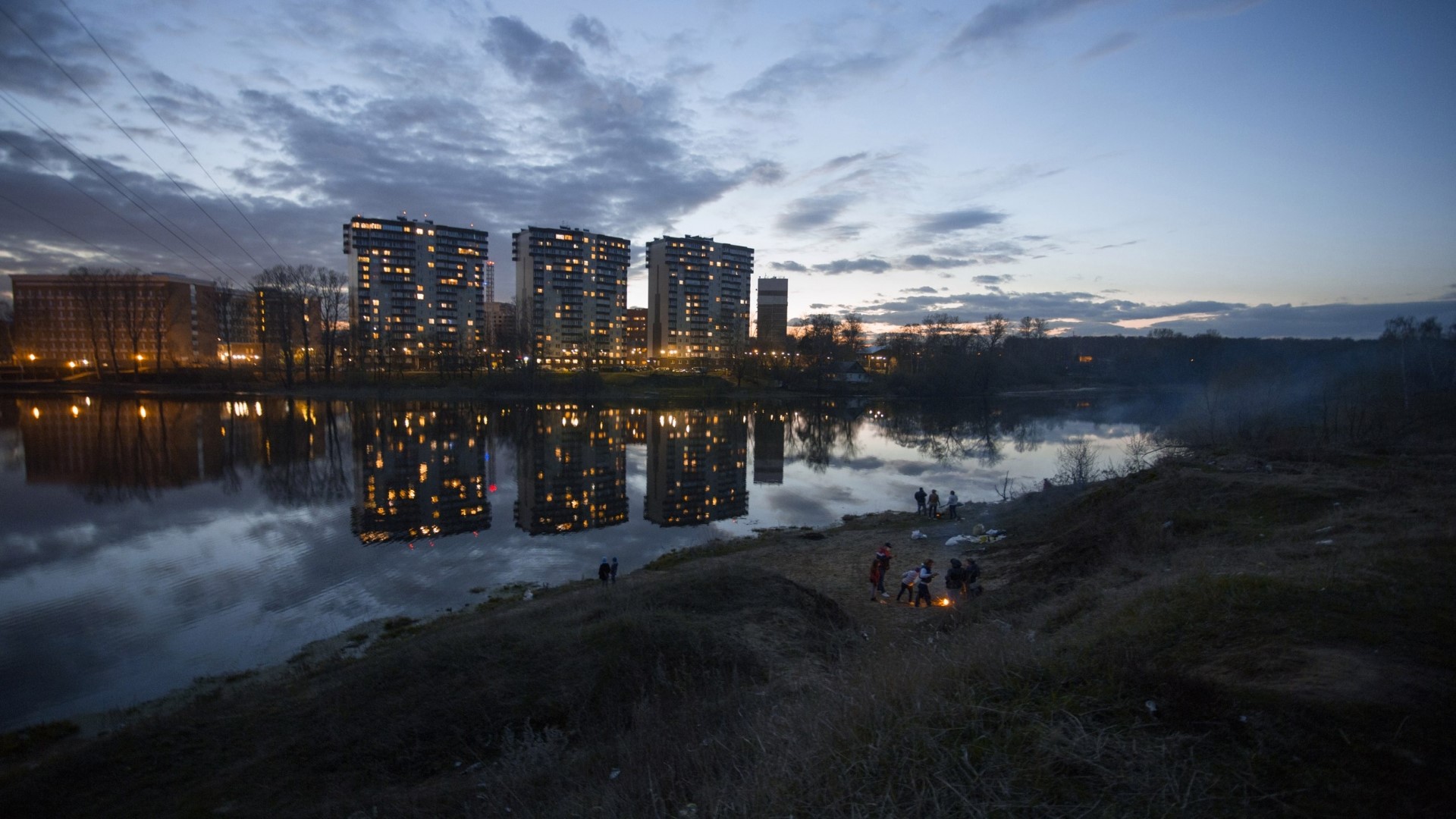 Apartment buildings reflected in a river at dusk with people by the shore HD Wallpaper