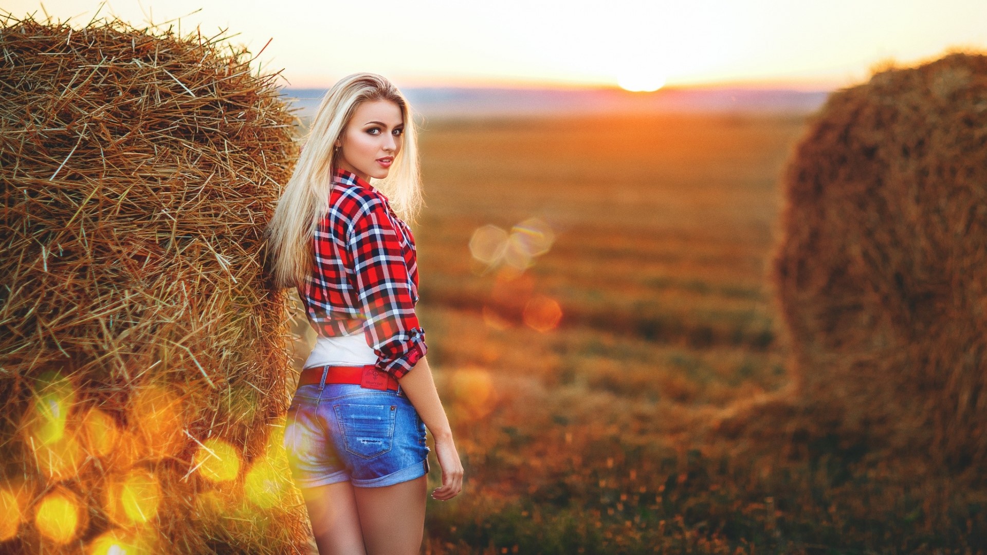 Woman in plaid shirt and denim shorts by hay bales at sunset HD Wallpaper