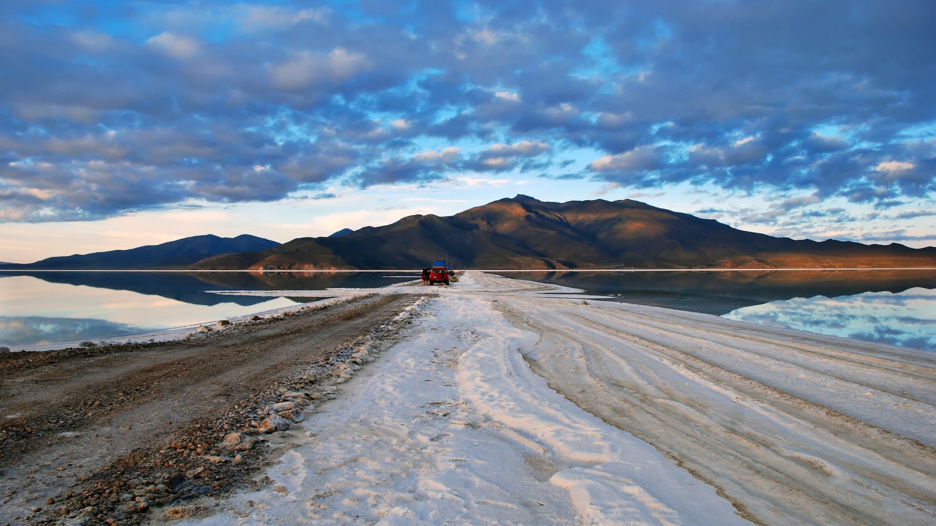 Red vehicle on a salt flat with mountains and clouds HD Wallpaper