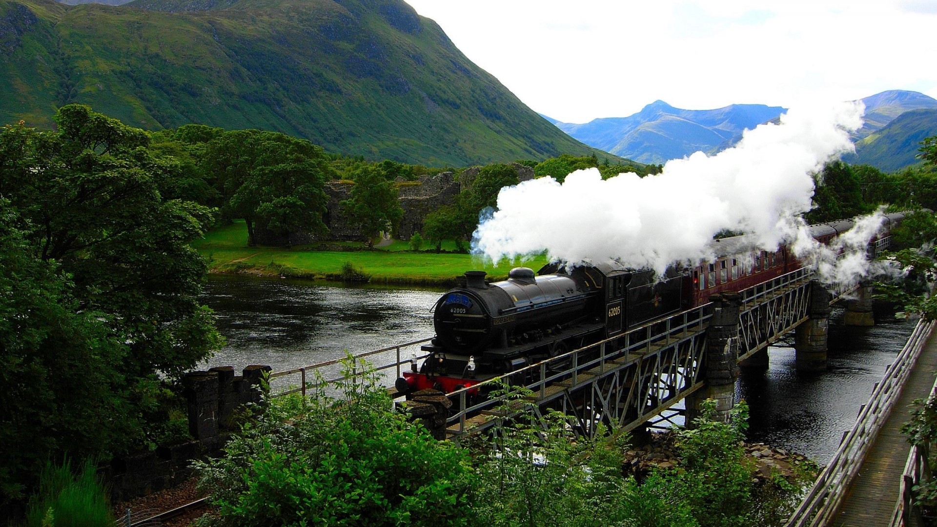Steam train crossing a bridge over a river in a mountainous landscape HD Wallpaper