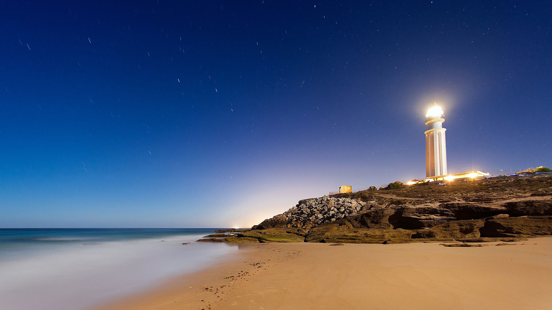 Lighthouse on a rocky coast at night HD Wallpaper