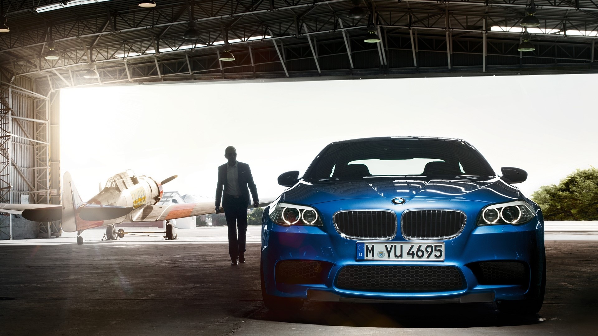 Blue BMW M5 parked in a hangar with a vintage airplane HD Wallpaper