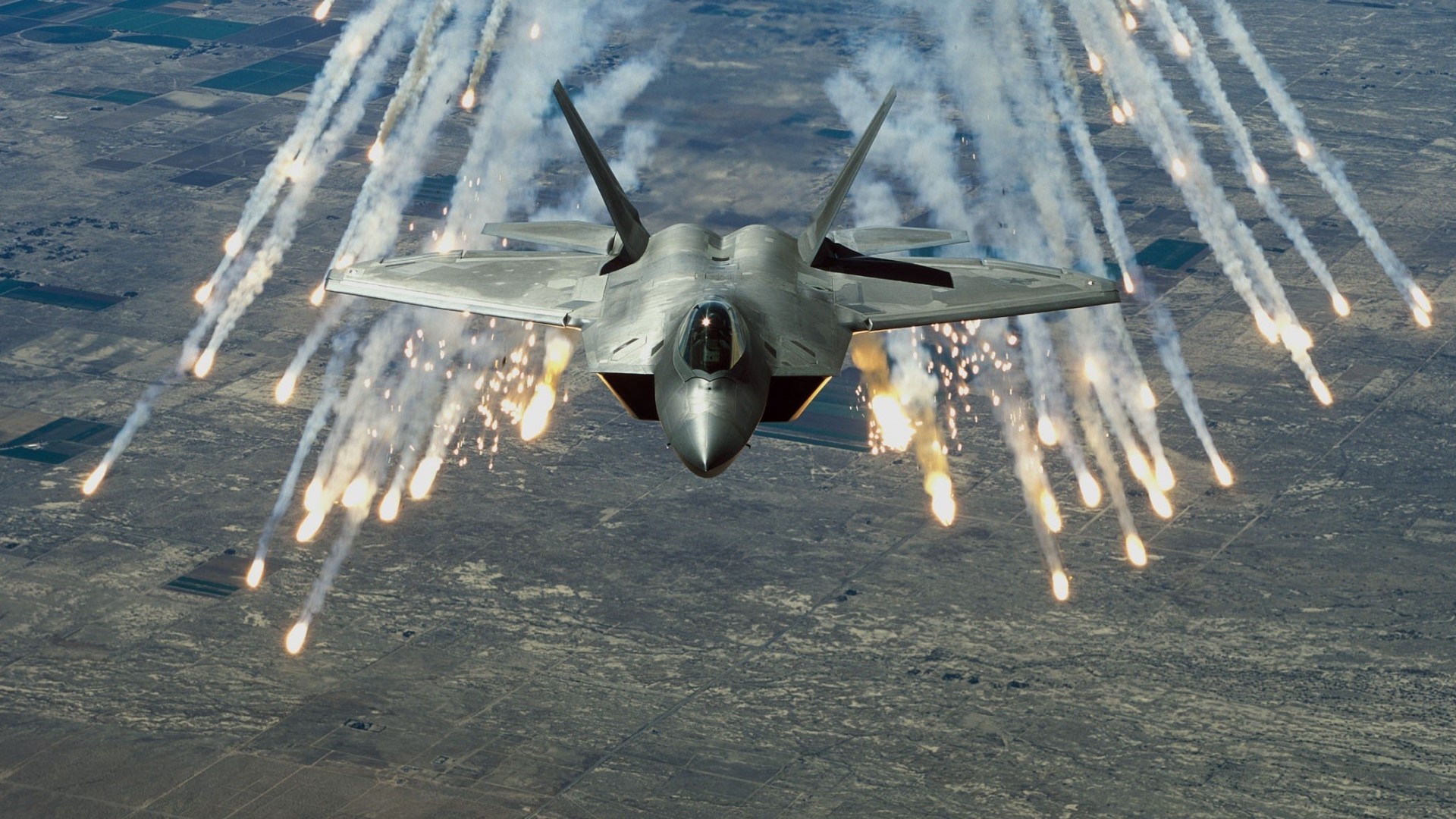 F-22 Raptor releasing flares over desert landscape HD Wallpaper