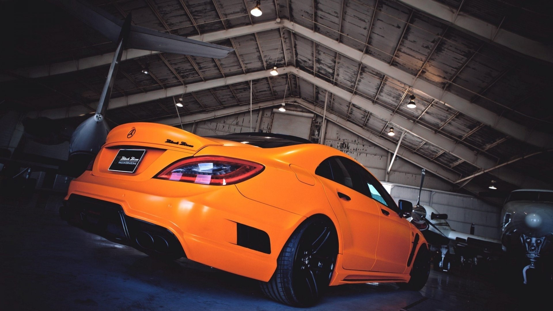 Orange Mercedes-Benz CLS in a hangar with airplanes HD Wallpaper