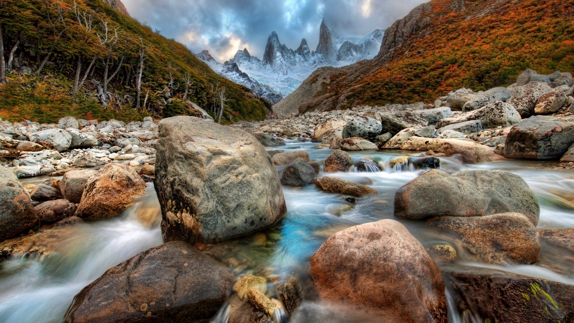 Mountain river flowing through rocky terrain with autumn foliage and snow-capped peaks HD Wallpaper