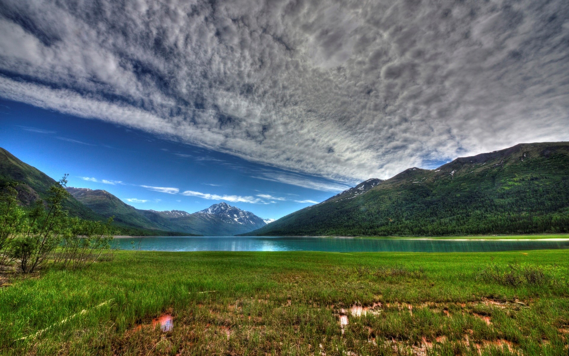 Grassy field with lake and mountains under cloudy sky HD Wallpaper