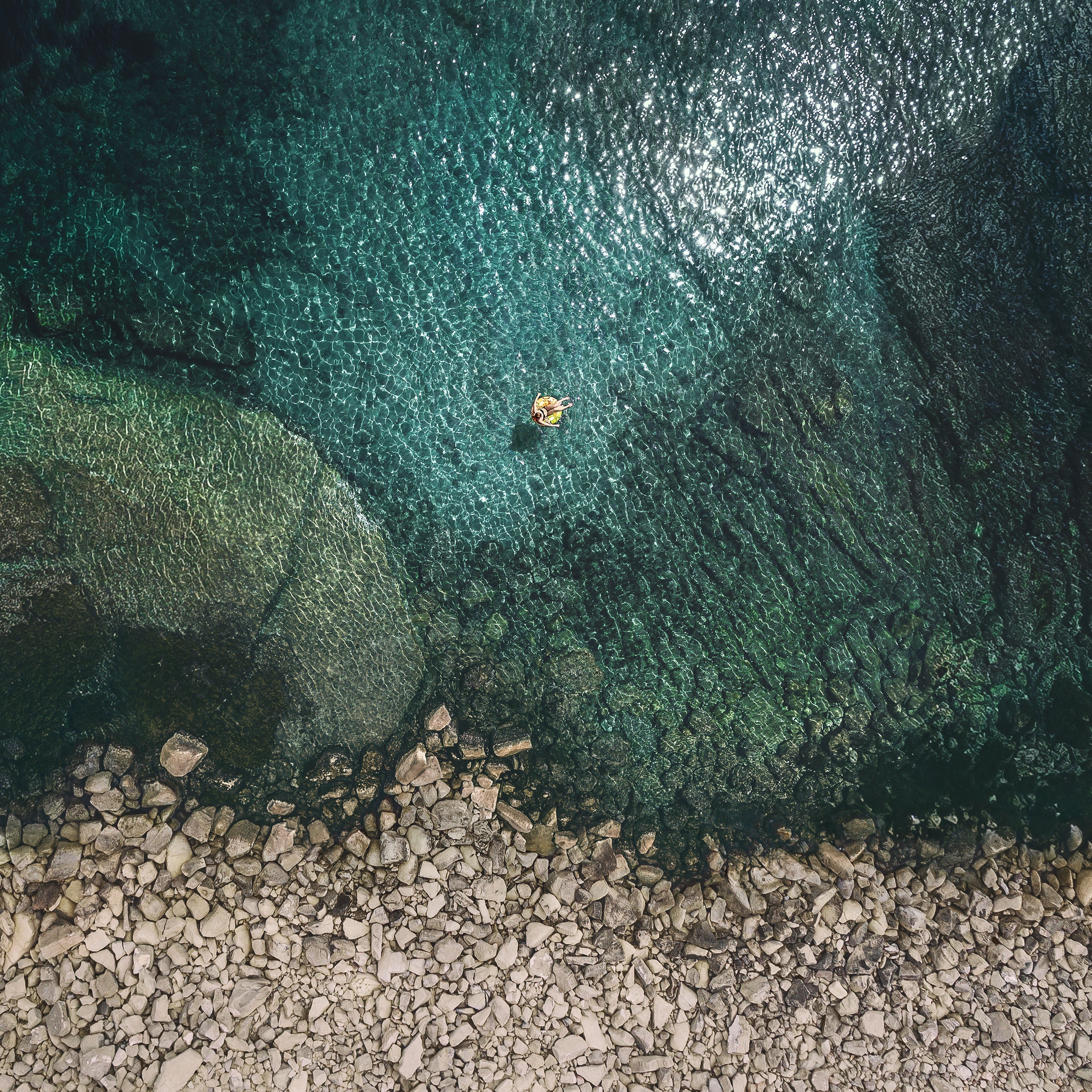 Person floating on inflatable in clear turquoise water near rocky shore HD Wallpaper