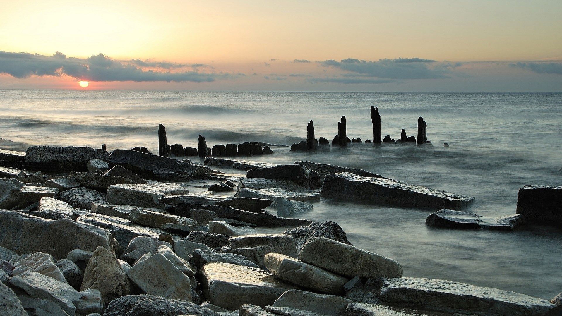 Rocky shoreline with wooden posts at sunrise HD Wallpaper