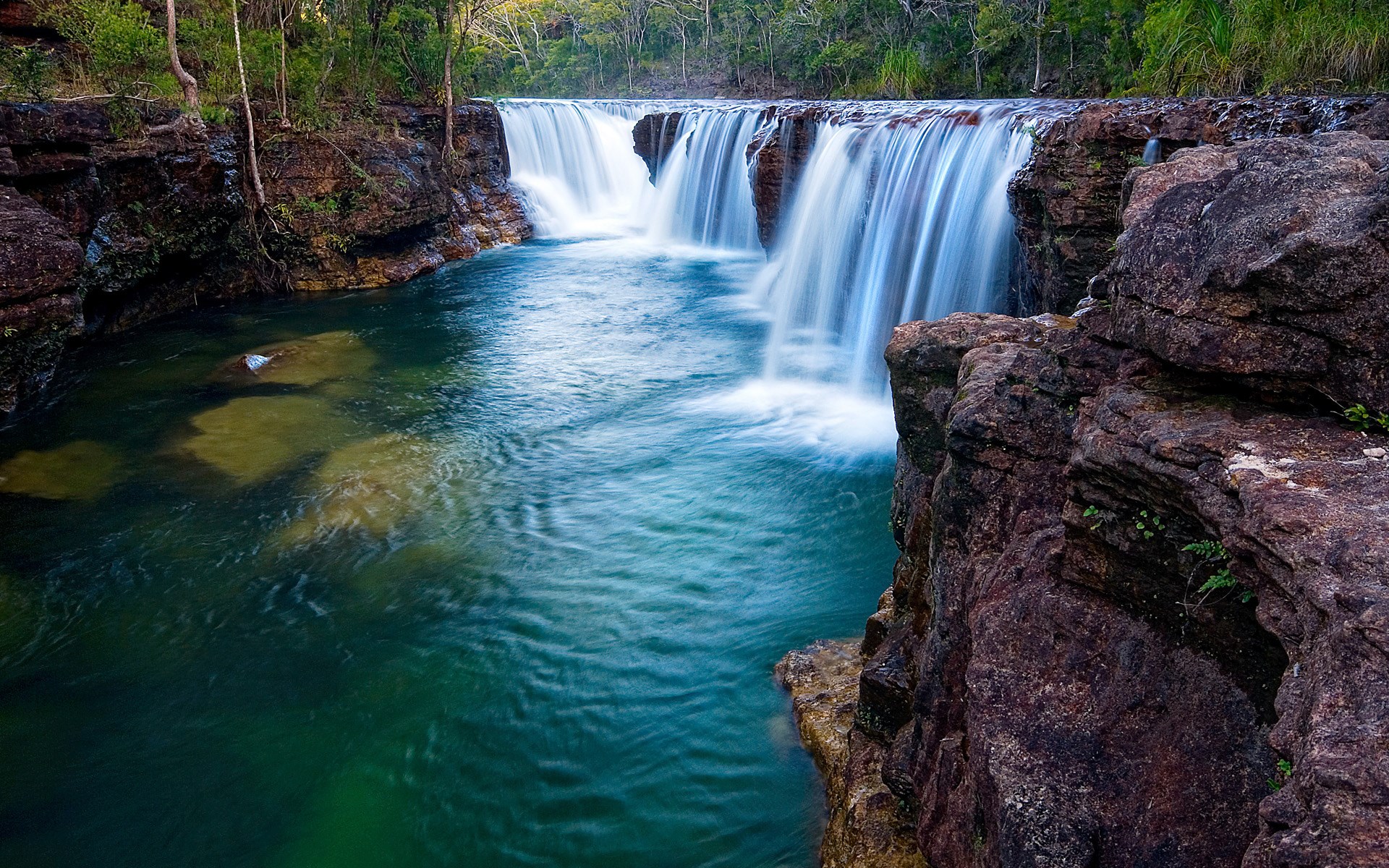 Waterfall cascading into a turquoise pool surrounded by rocks and trees HD Wallpaper