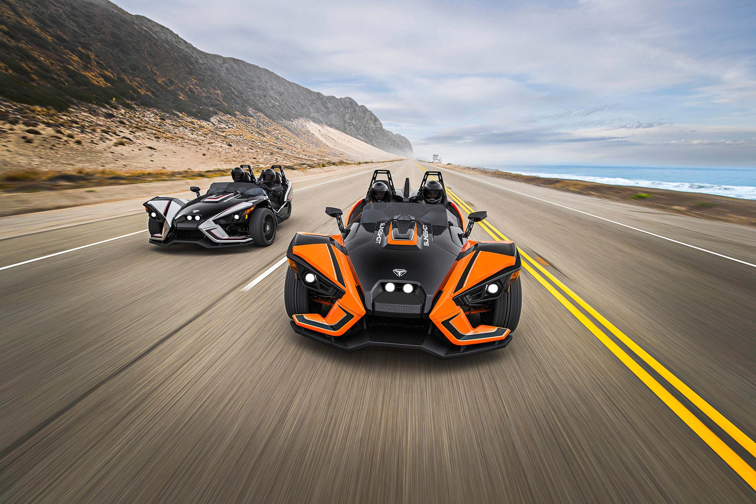 Two Polaris Slingshots driving on a coastal highway HD Wallpaper