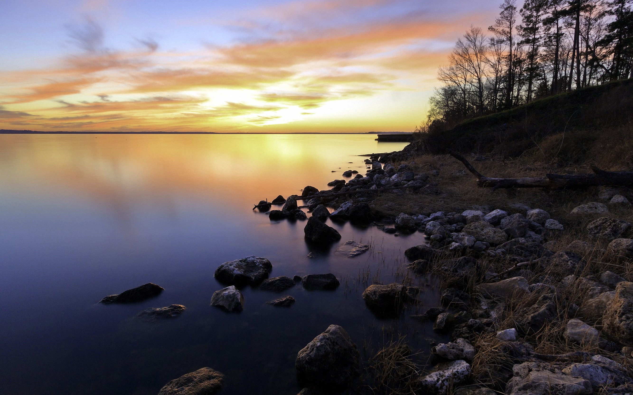Sunset over a calm lake with rocky shore and trees HD Wallpaper