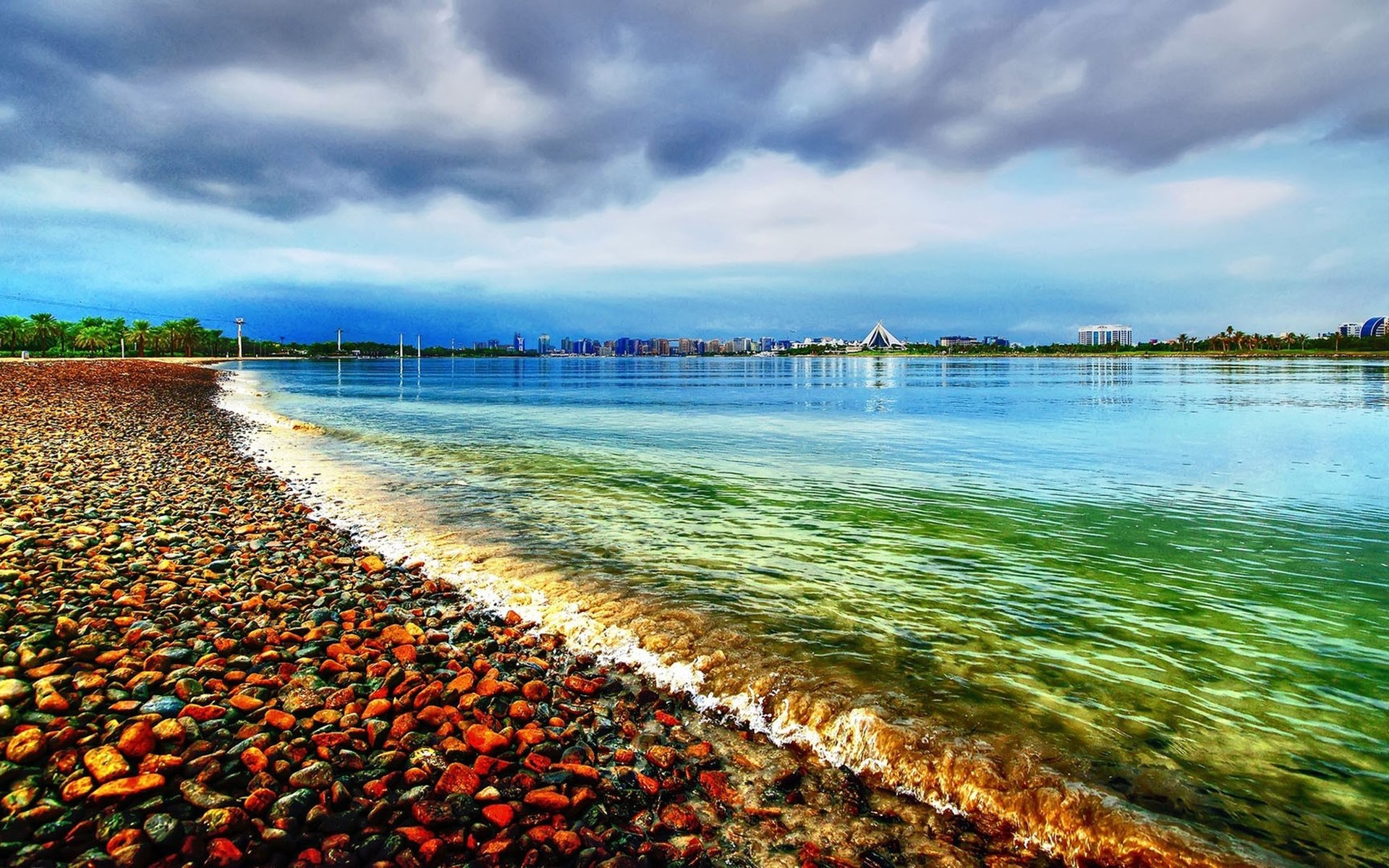 Pebble beach with calm water and city skyline HD Wallpaper