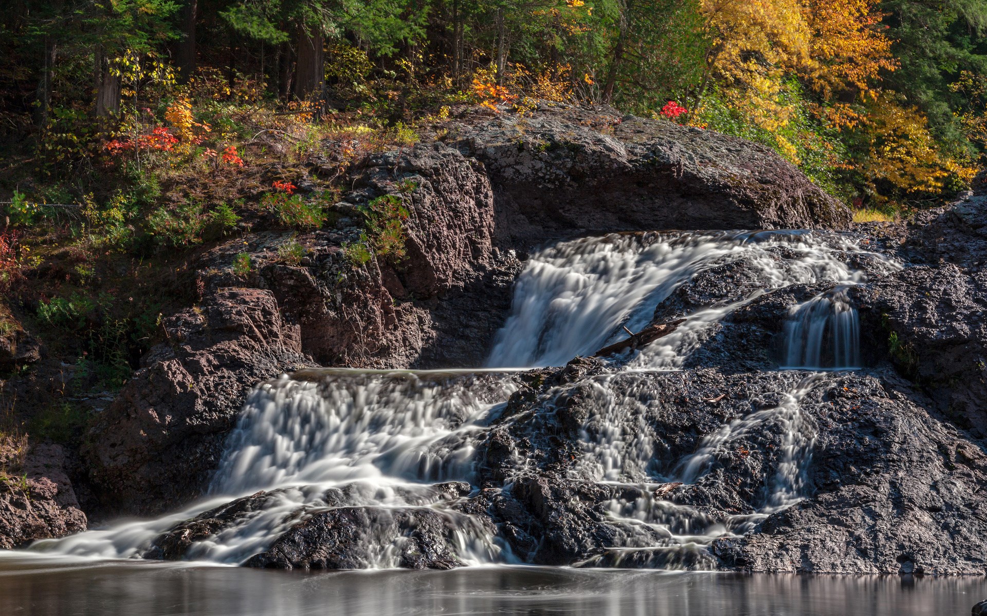 Waterfall cascading over dark rocks with autumn foliage HD Wallpaper
