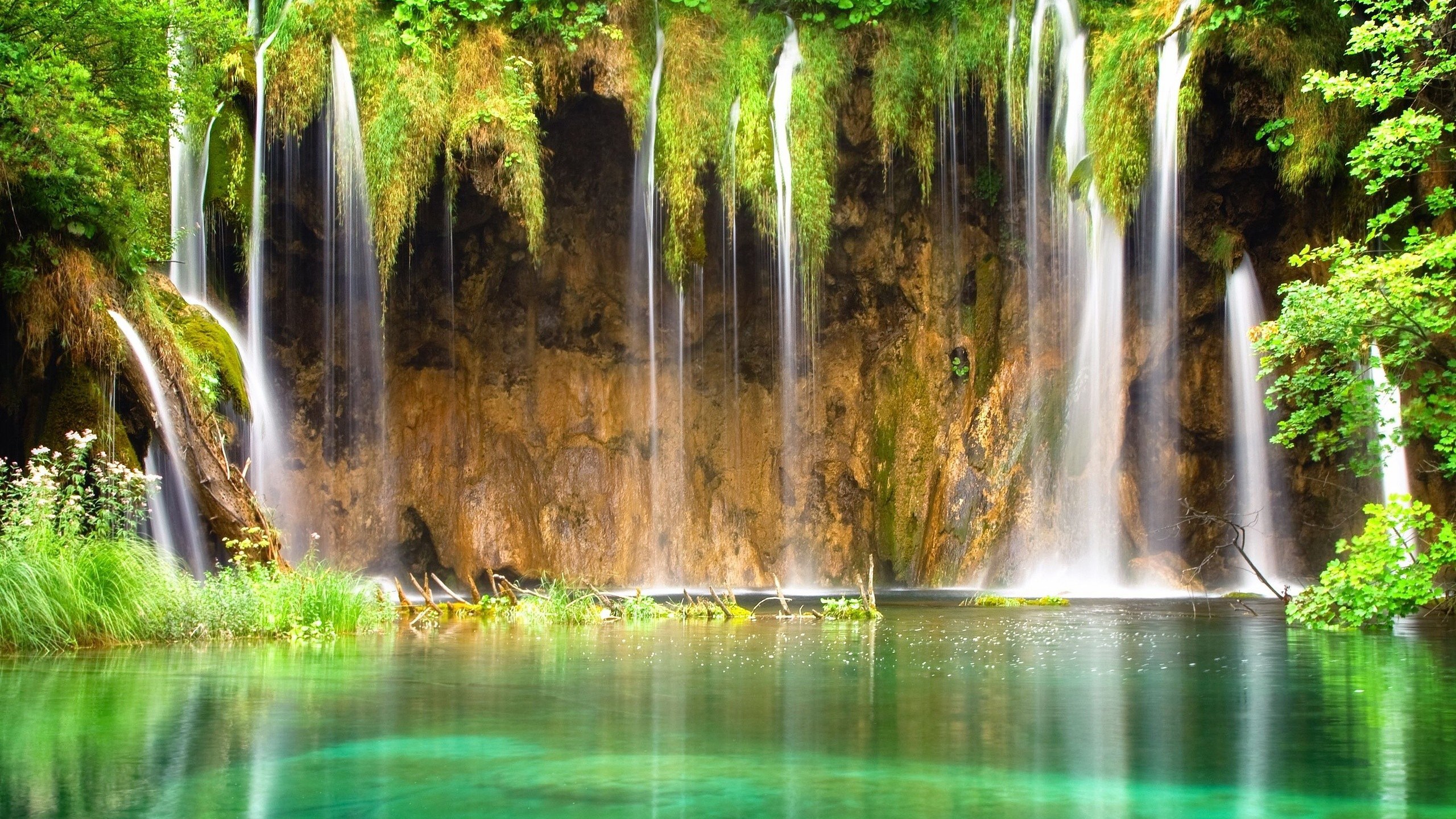 Waterfalls cascading over mossy rocks into a clear green lake HD Wallpaper