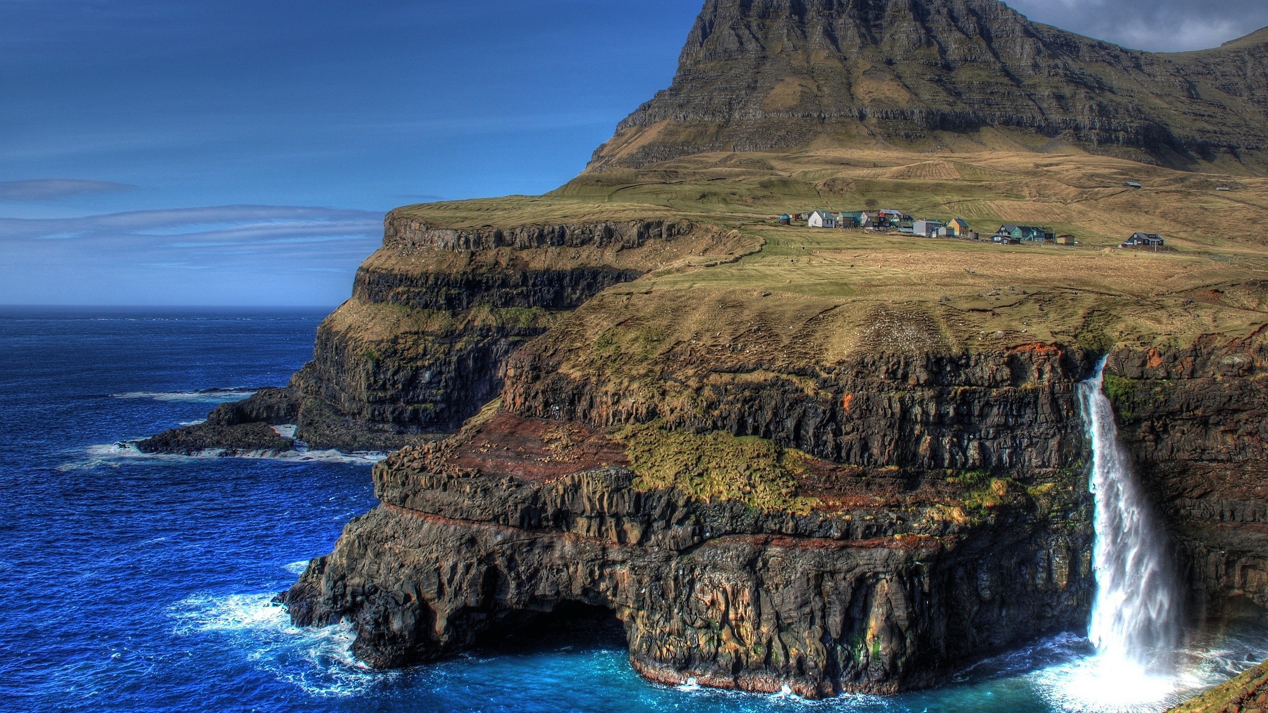 Waterfall cascading from cliffs into the ocean near a village HD Wallpaper