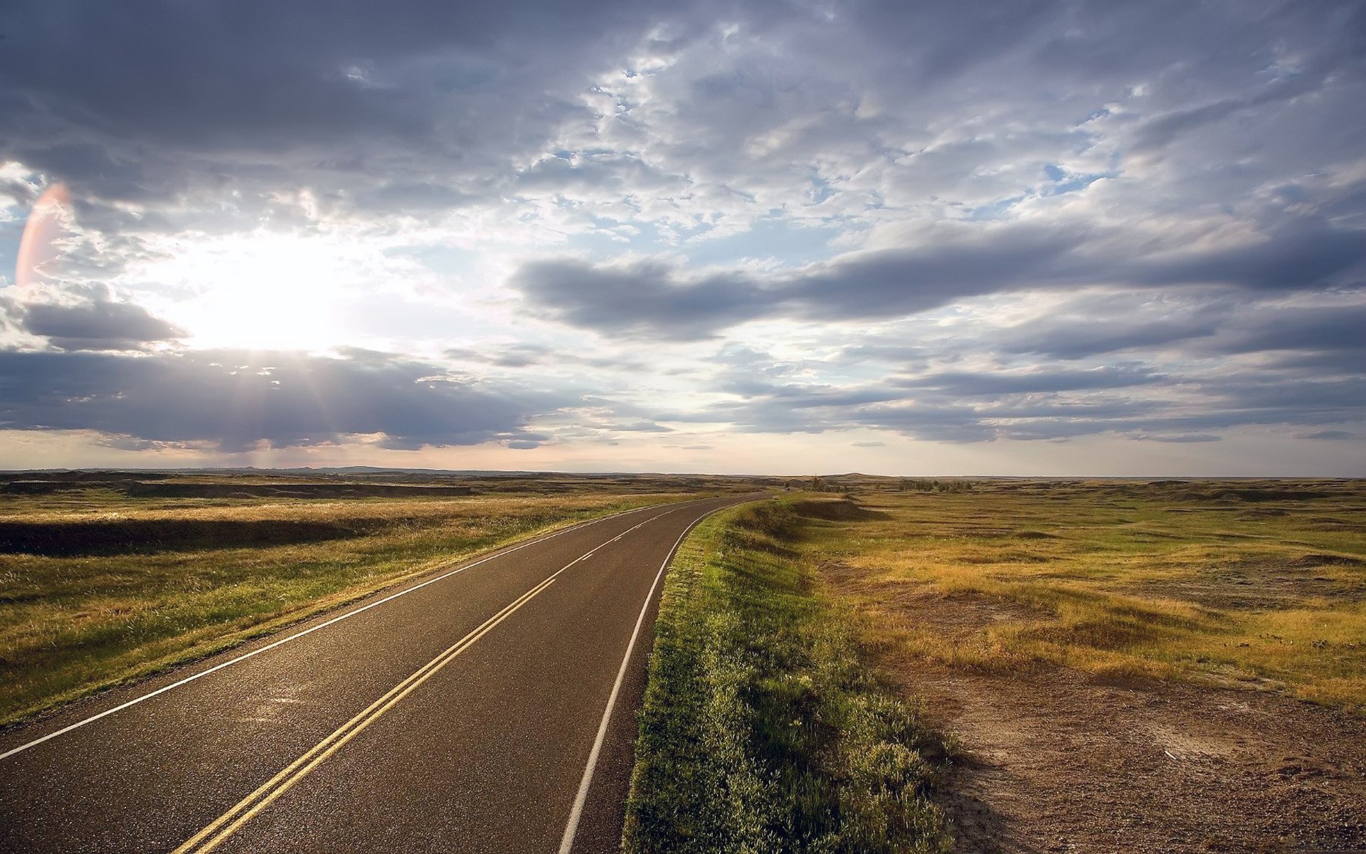 Road through a grassy plain under a cloudy sky HD Wallpaper
